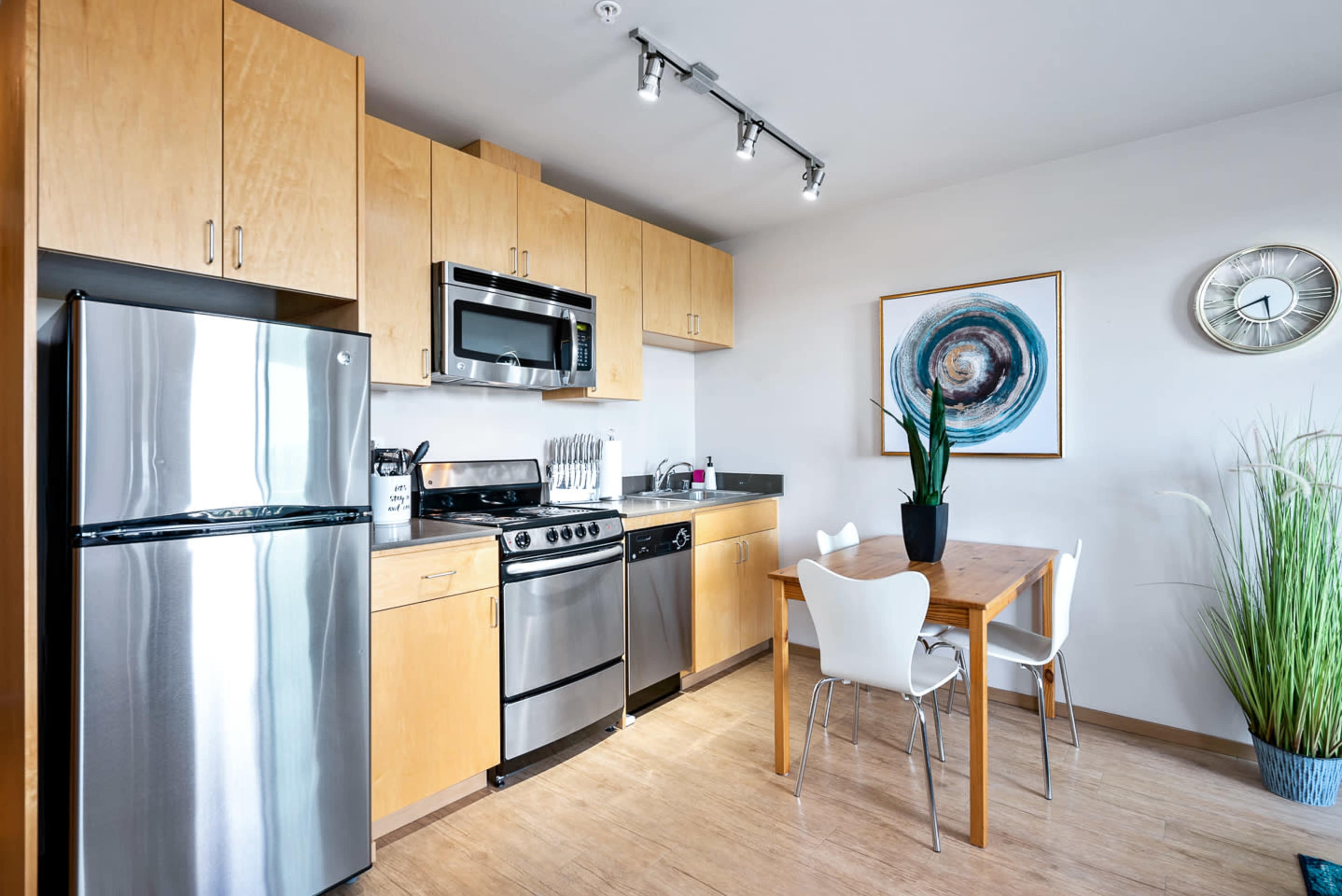 The image shows a modern kitchen area featuring stainless steel appliances, wooden cabinets, a dining table with white chairs, and a decorative plant.