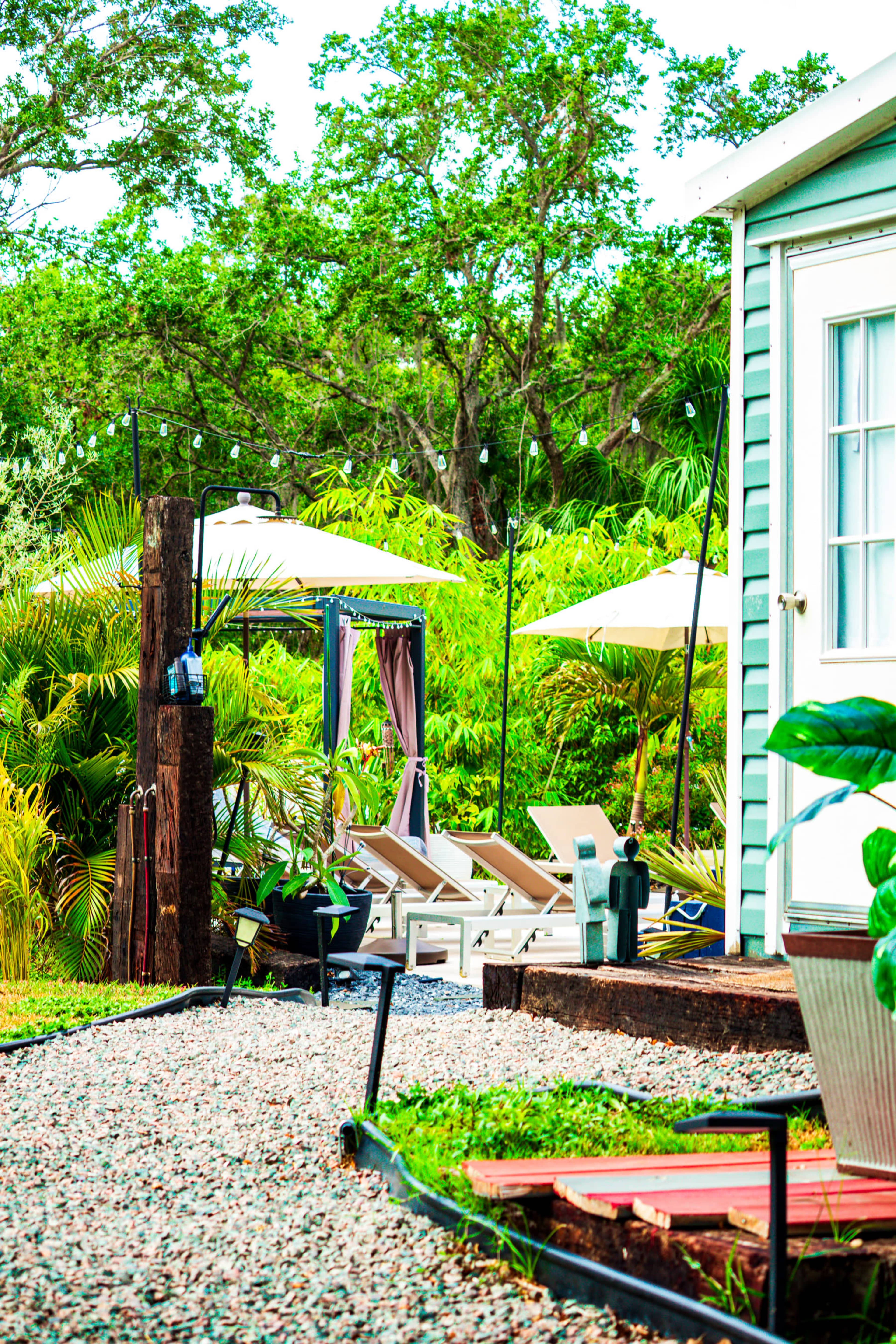A pathway of gravel leads to lounge chairs under umbrellas beside a small green building surrounded by lush greenery.