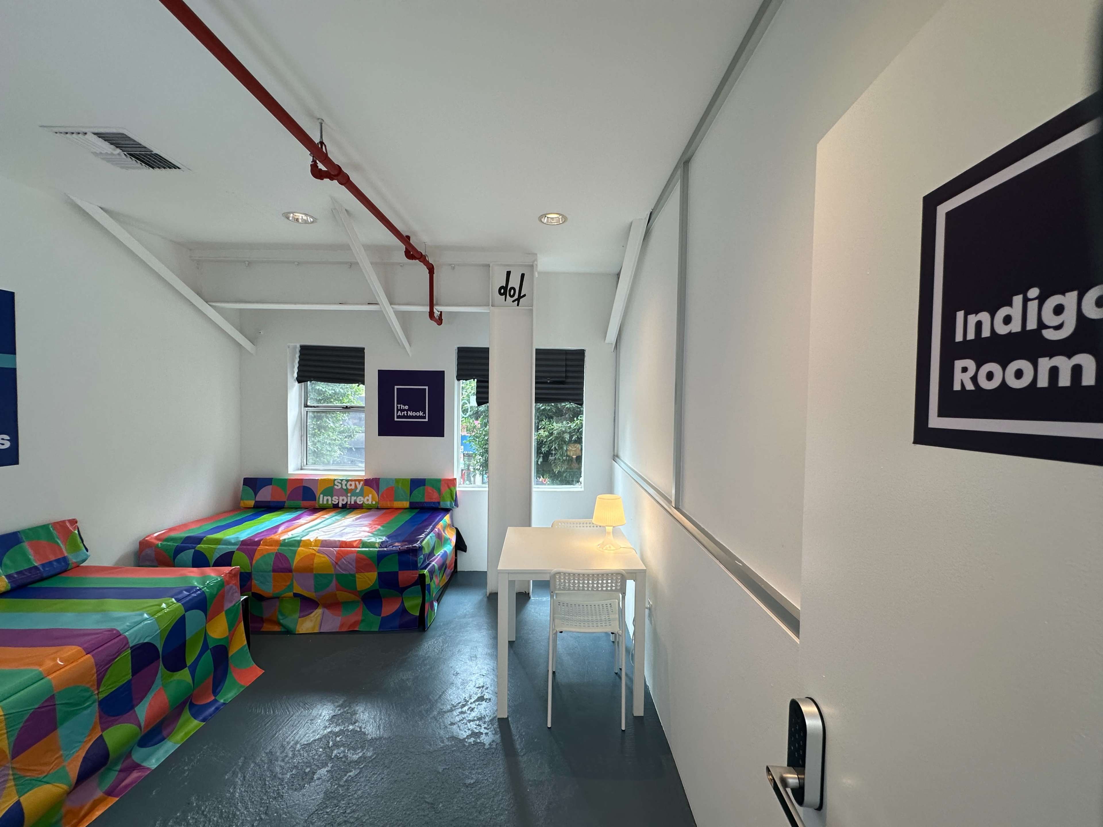 The image shows a brightly colored room with patterned beddings, a small white table, and large windows.