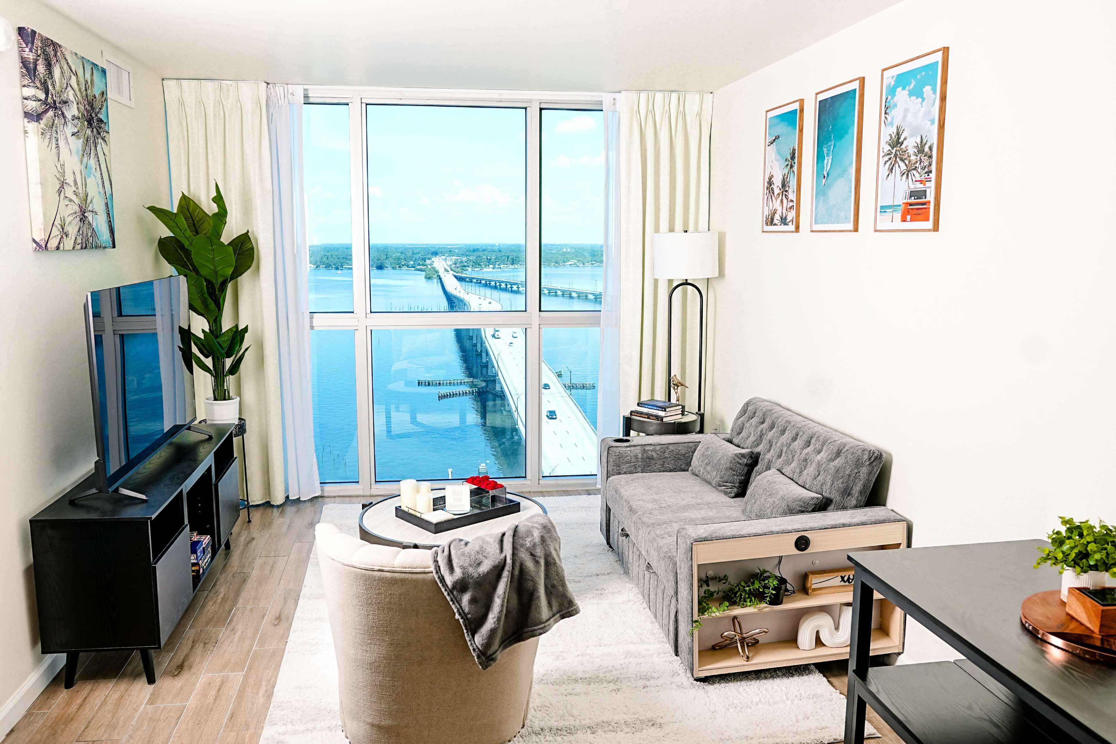 A modern living room features a grey sofa, a round chair, a television unit, and large windows offering a view of a bridge over water.