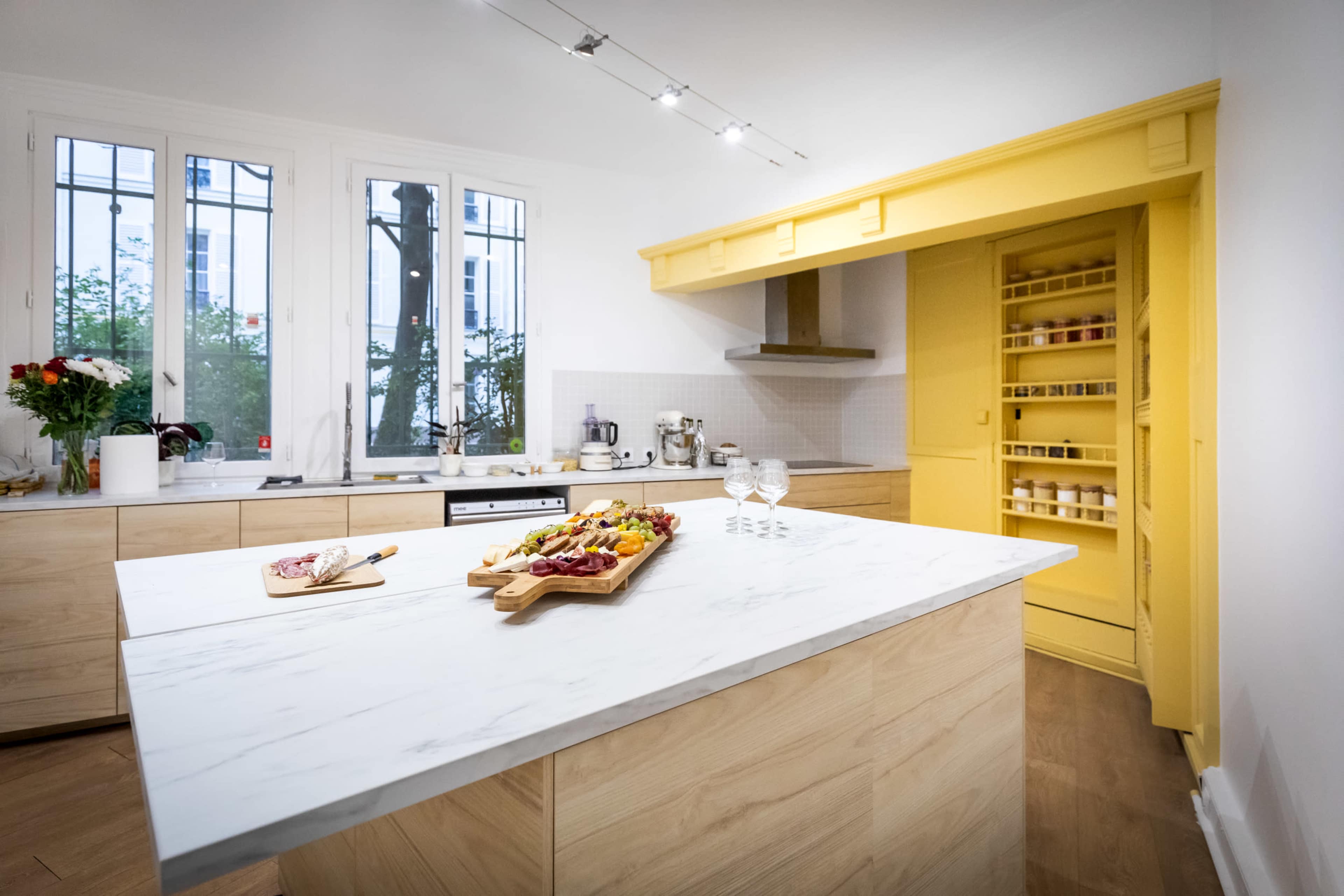 A modern kitchen features a marble island with a charcuterie board, wooden cabinetry, and a yellow pantry against a bright, clean backdrop with large windows.