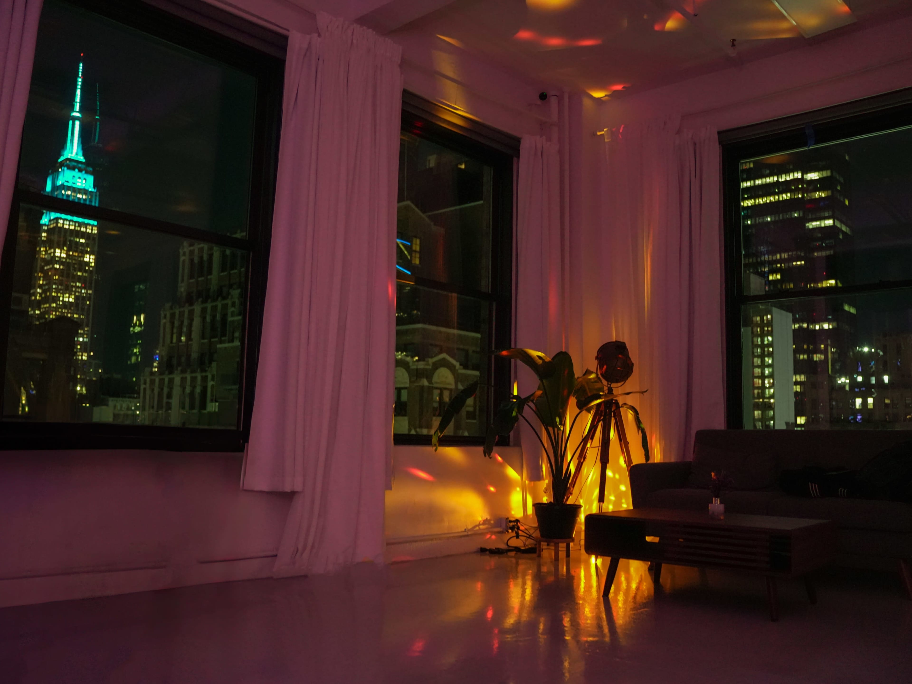 A brightly lit room features large windows with a view of the Empire State Building, accompanied by a lounge area with a plant and a spotlight.