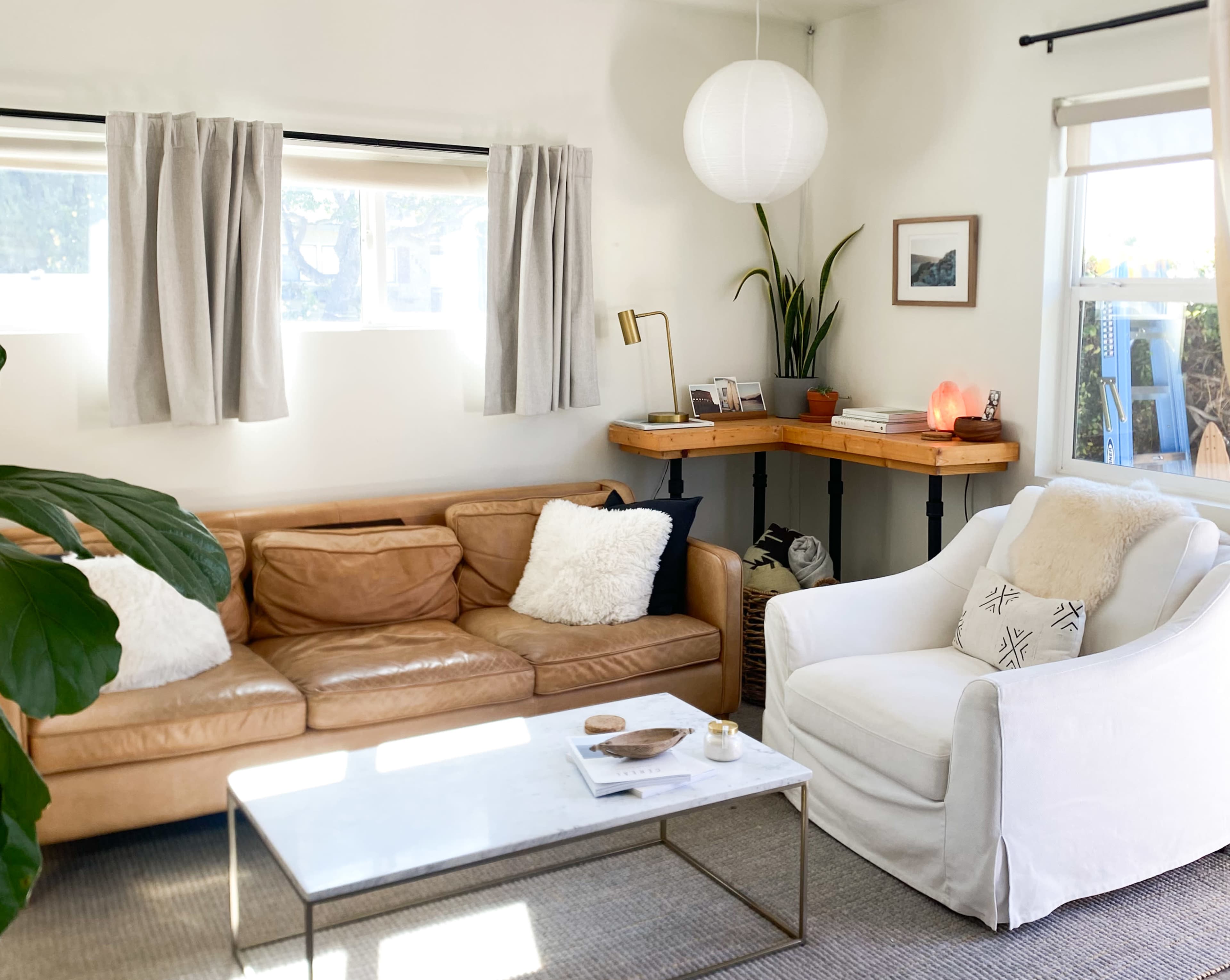 The scene features a cozy living room with a light-colored sofa, a white armchair, a coffee table, and a desk, complemented by natural light from the windows.