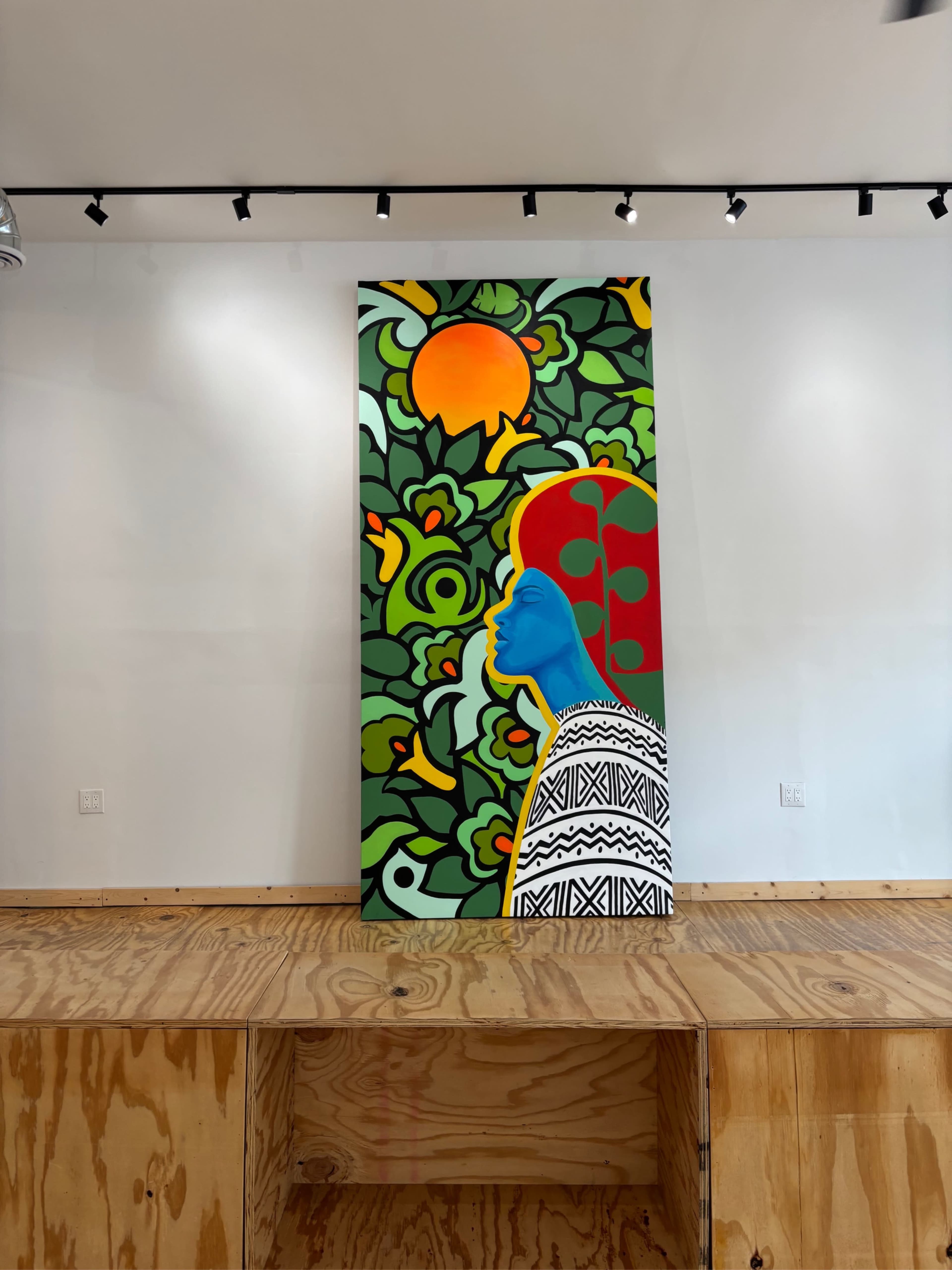 The image shows a large, colorful mural depicting a blue-faced figure surrounded by vibrant greenery and a sun on a white wall.