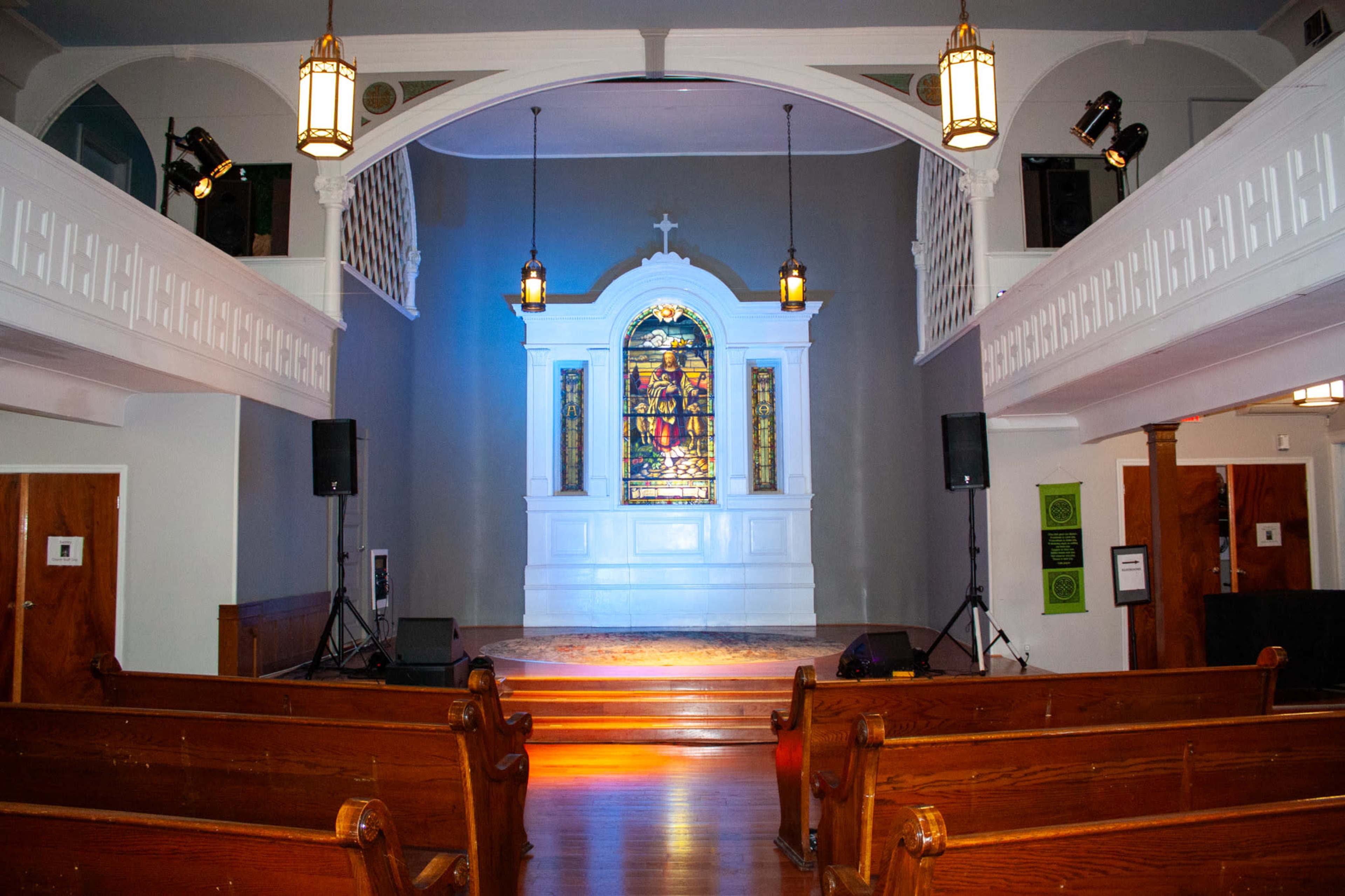 The image shows the interior of a church featuring a stained glass window with a stage area, wooden pews, and soft lighting.