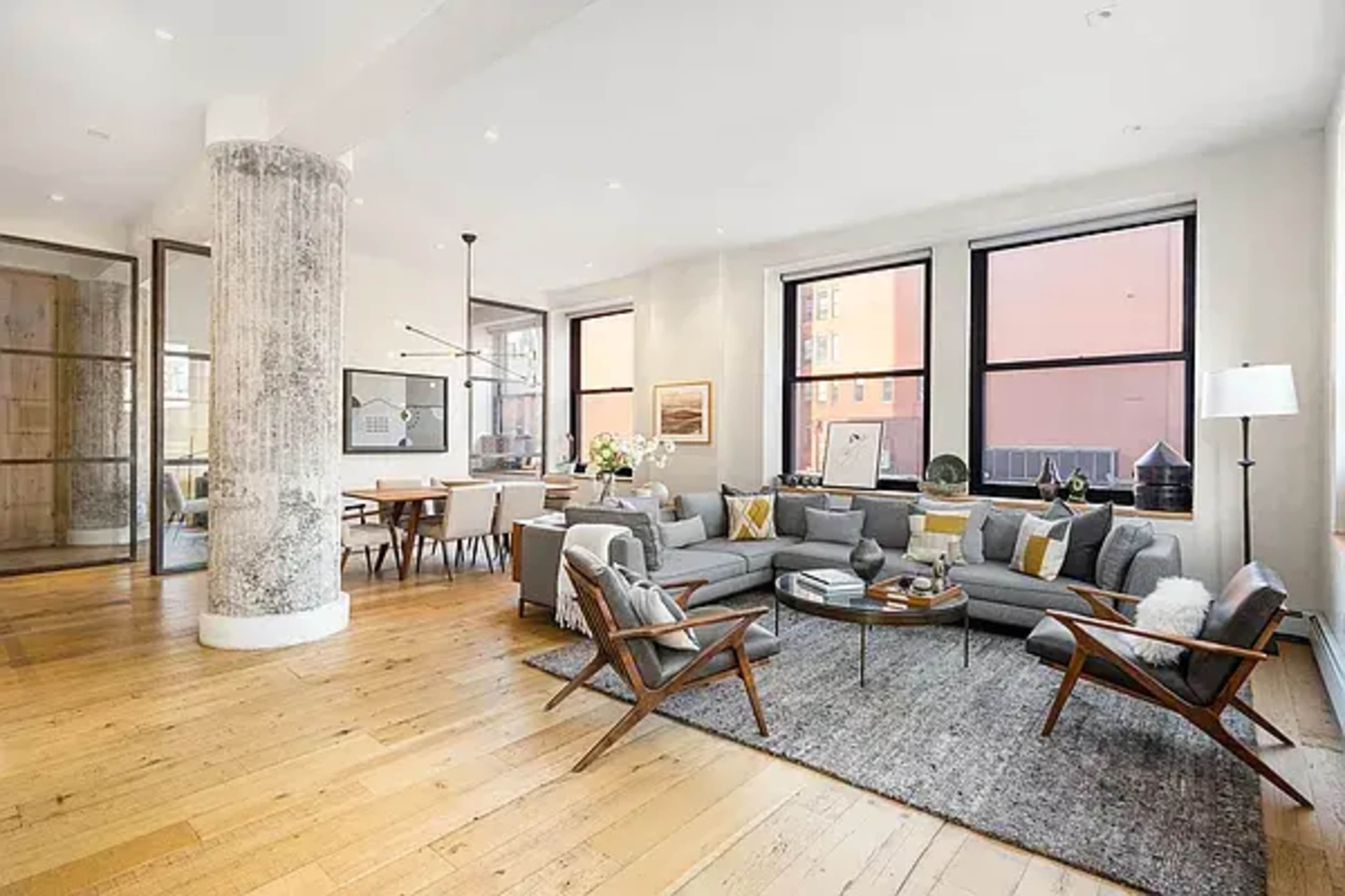 The image depicts a modern, open-concept living space featuring a gray sectional sofa, a coffee table, and large windows with natural light illuminating the wooden floor.
