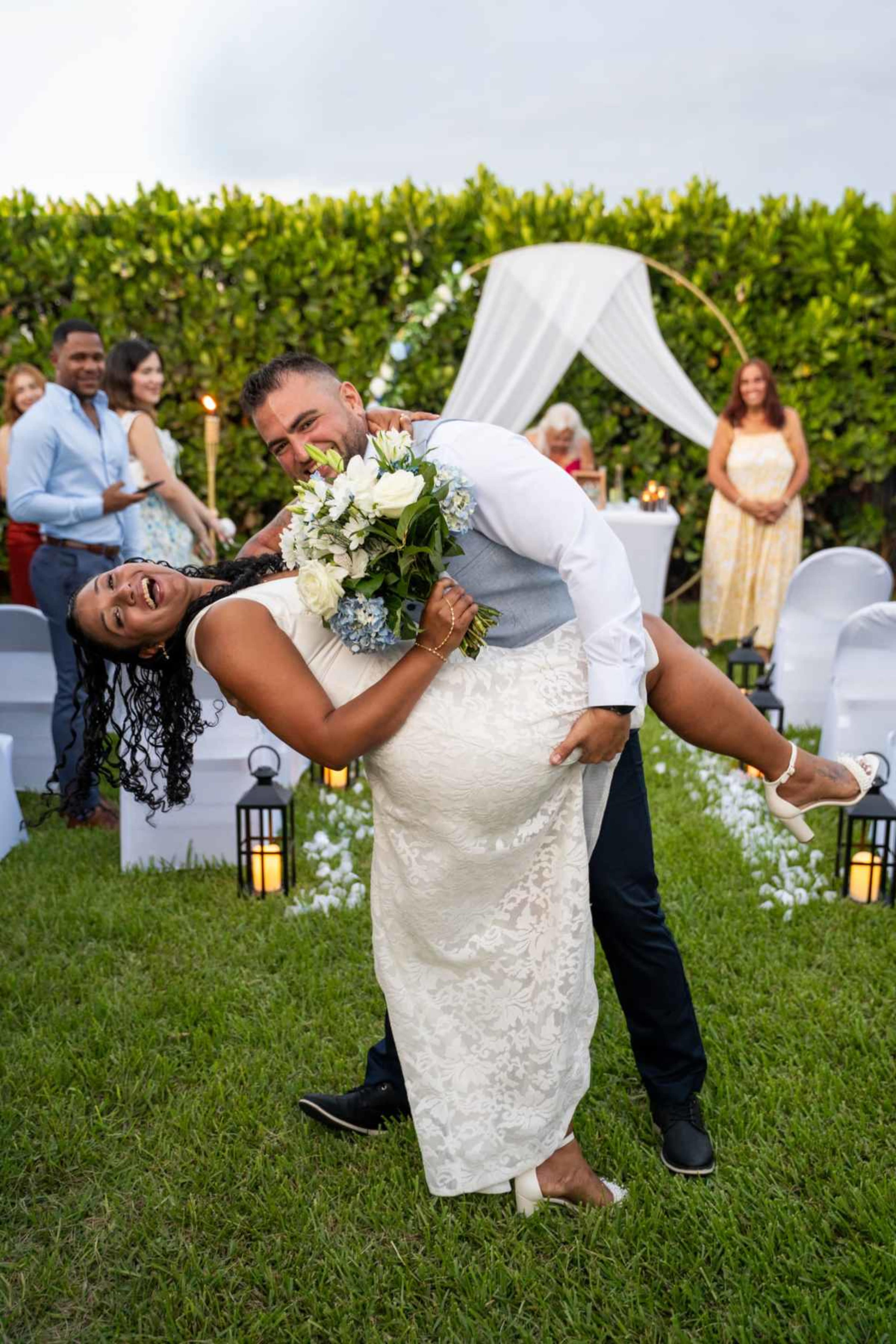 A groom swings his bride in a playful dip at an outdoor wedding ceremony surrounded by guests and decorative elements.
