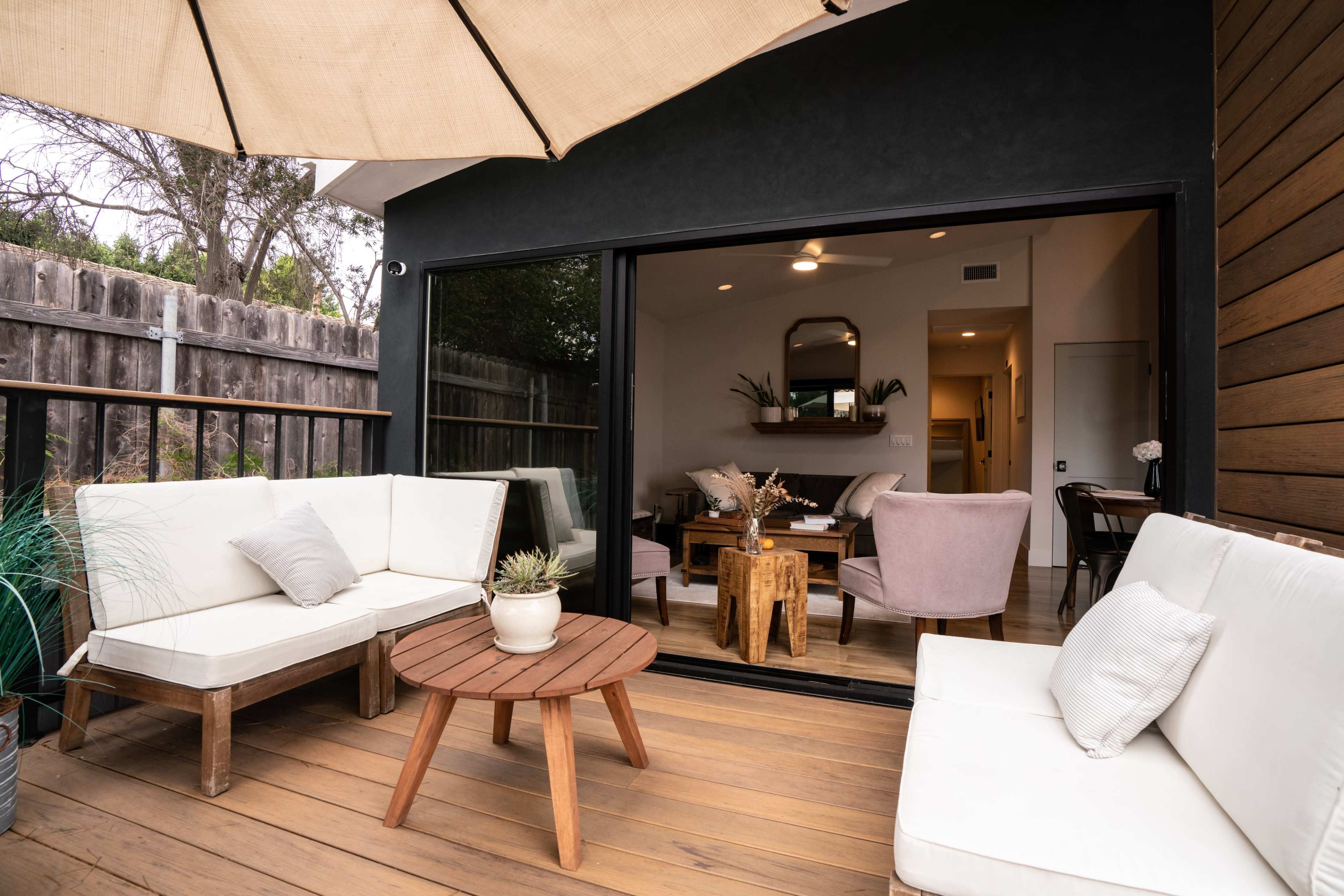 A modern patio features a seating area with a wooden coffee table, an umbrella, and a view into a well-lit interior space.