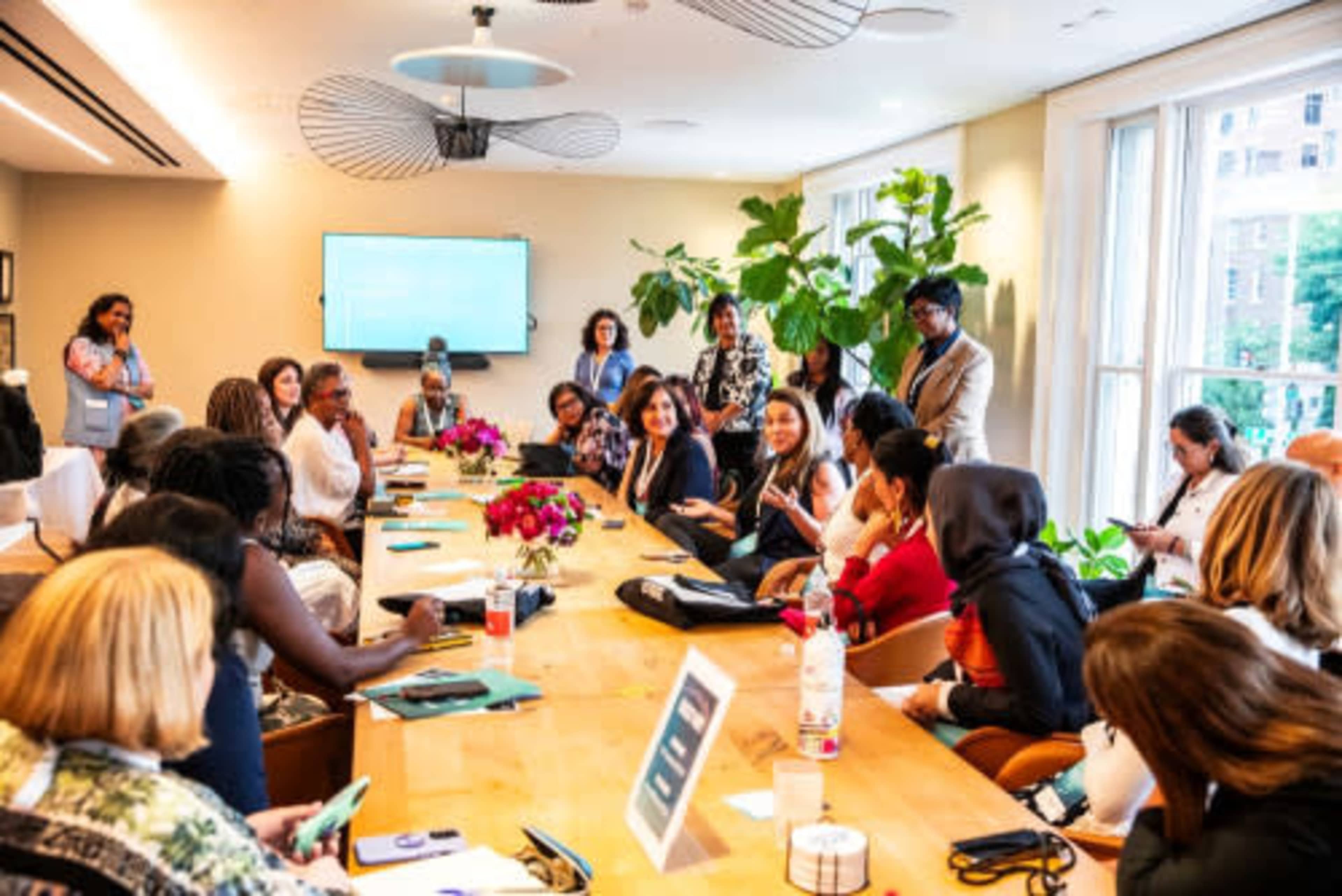 A diverse group of people is engaged in discussion around a long table in a well-lit conference room adorned with plants and a large screen.