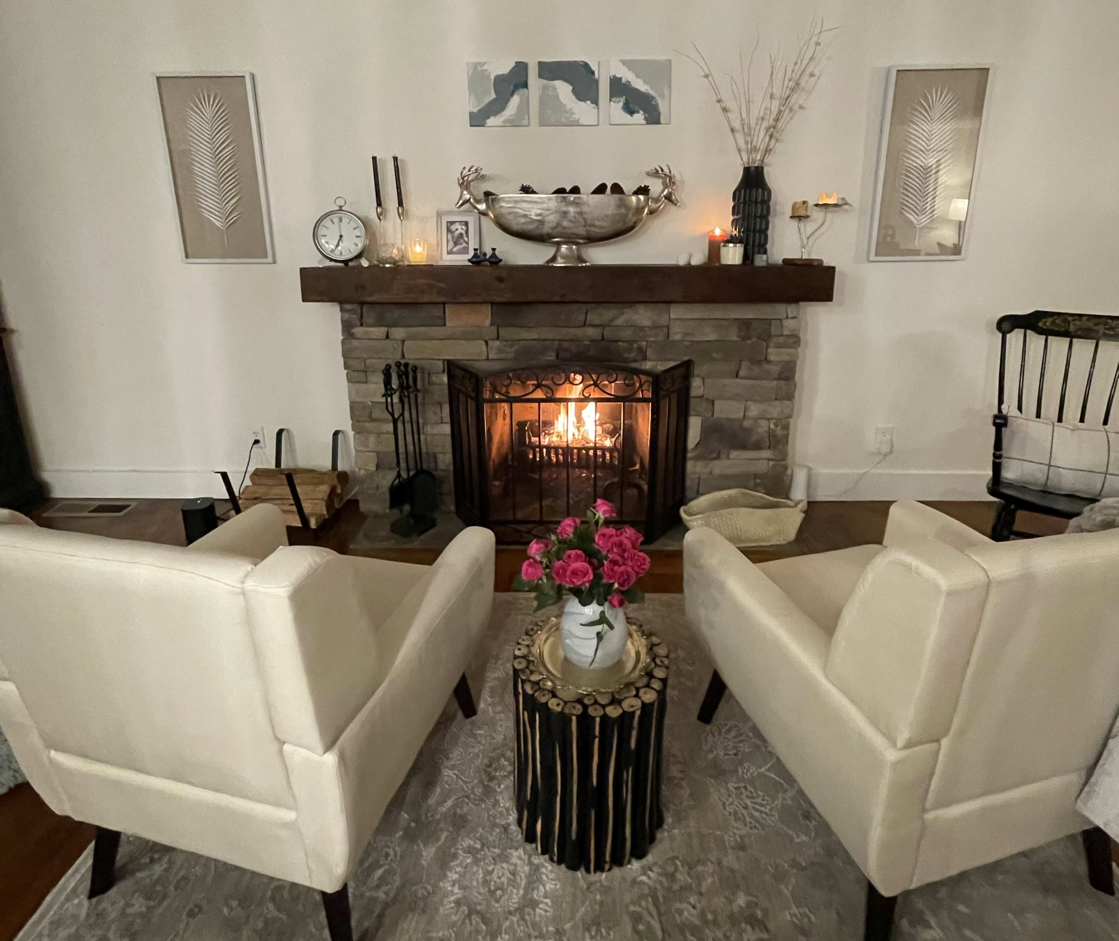 Two white upholstered chairs face a stone fireplace with a lit fire, surrounded by decor including framed art, a clock, and a floral centerpiece on a wooden stump table.