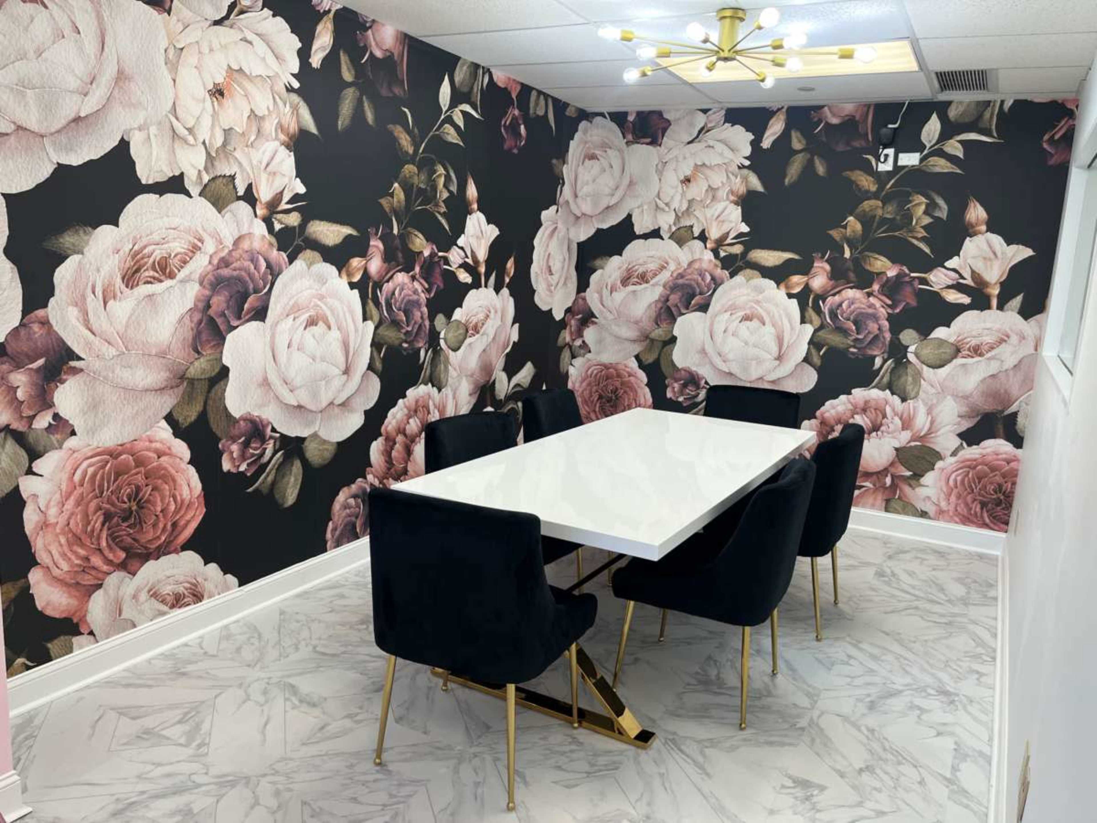 A conference room with a large white table surrounded by four black chairs, against a wall decorated with a floral wallpaper featuring large pink and white roses.
