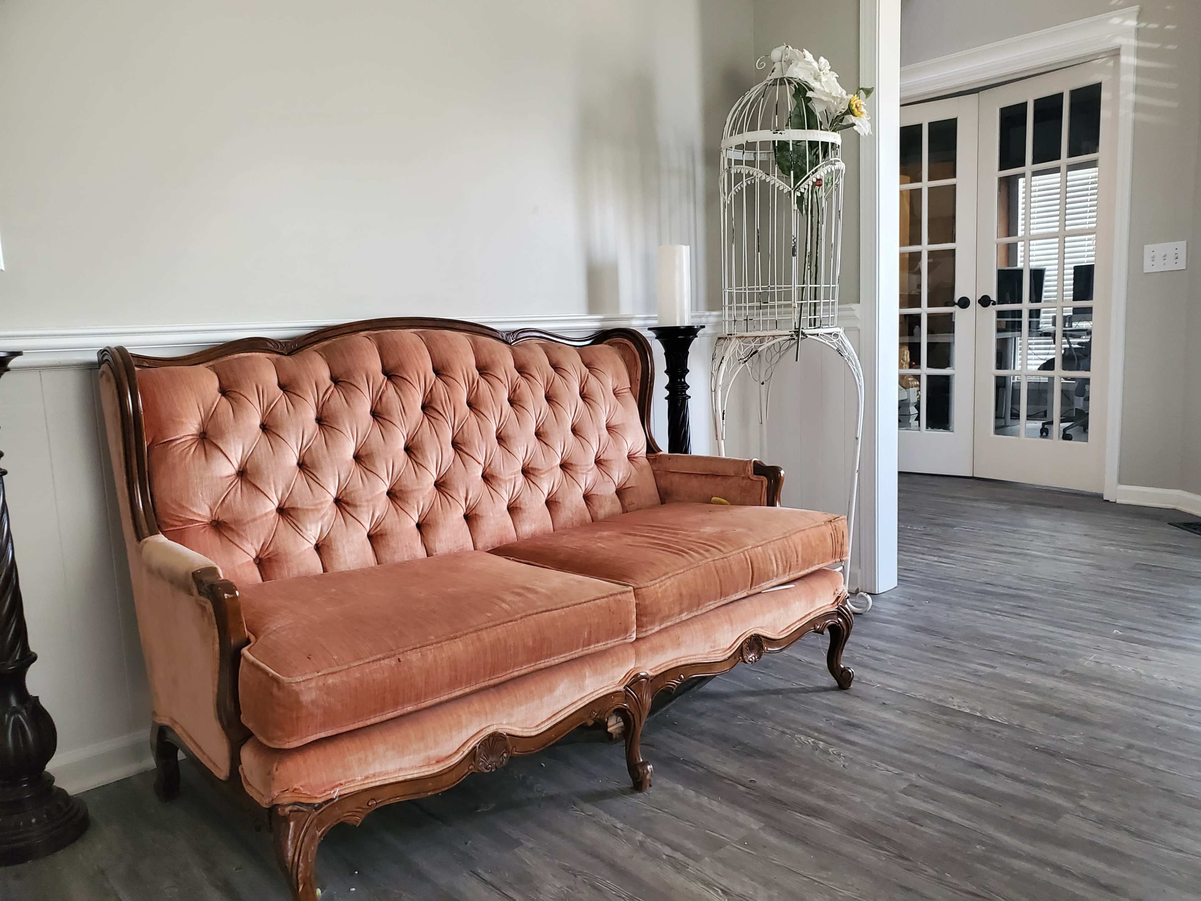 A vintage pink tufted sofa is positioned against a gray wall, with a white birdcage and a set of glass doors visible in the background.