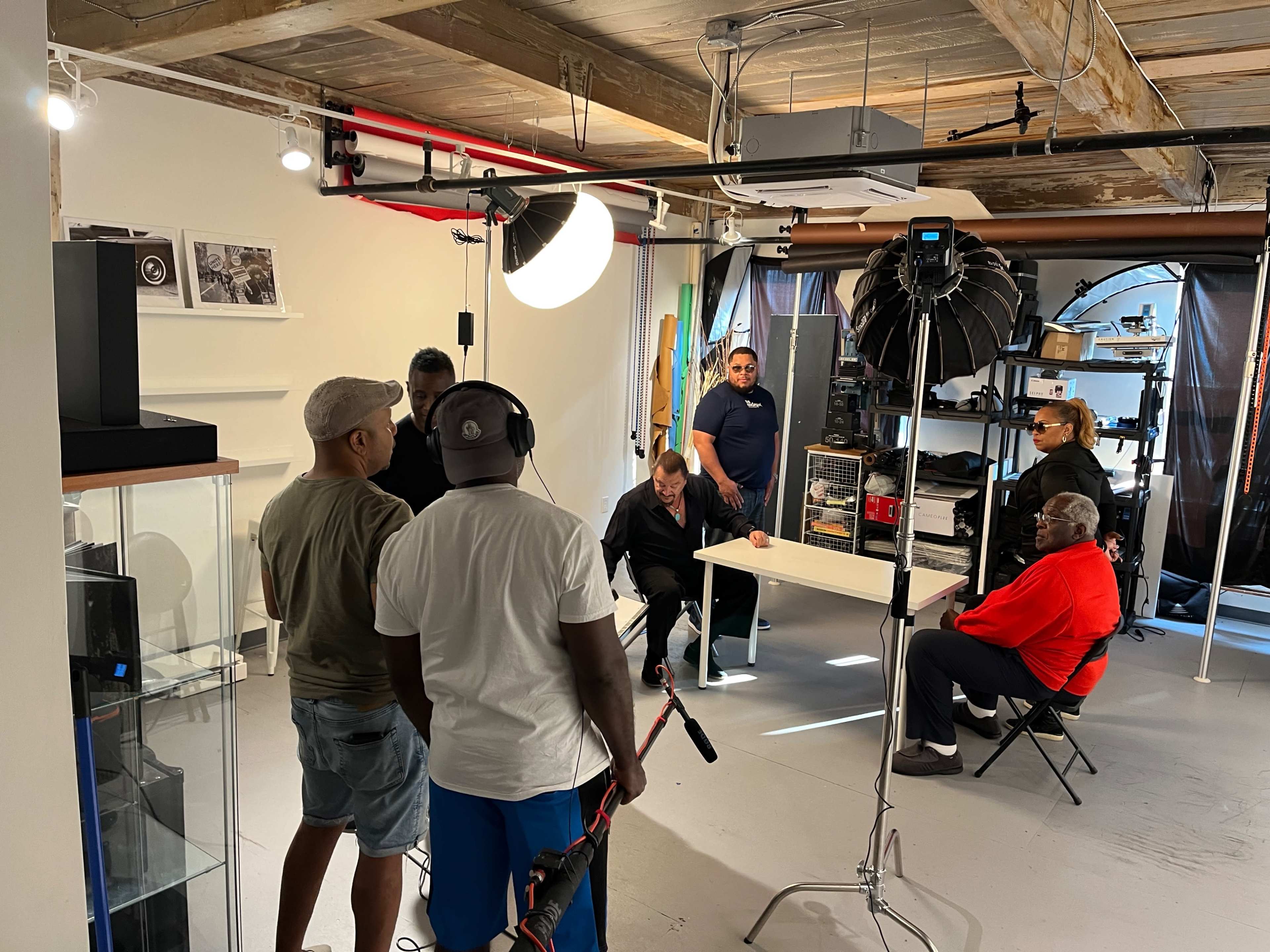 A group of five individuals engages in discussion around a camera setup in a studio space with shelves and lighting equipment.