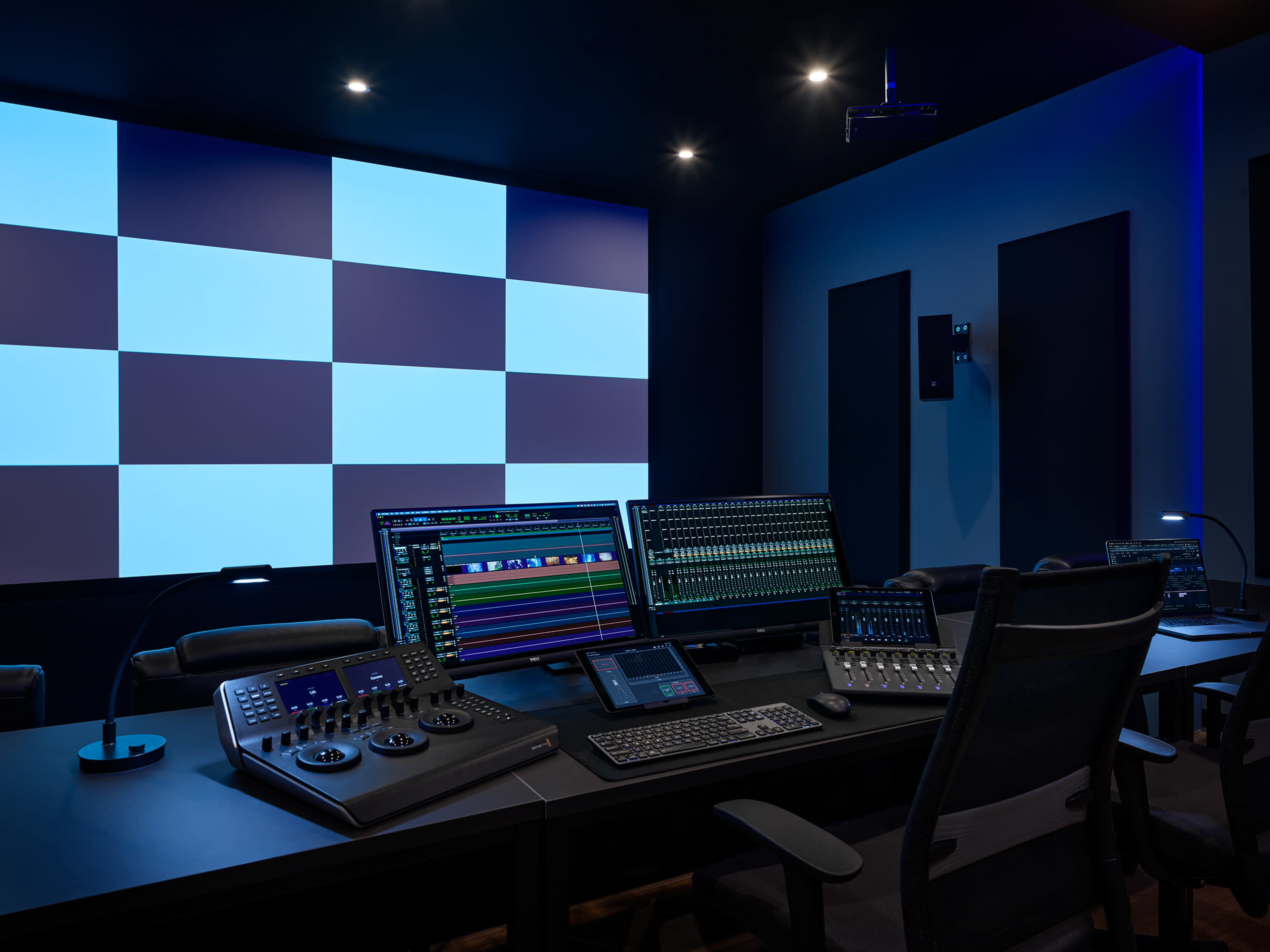 A modern audio-visual control room with multiple computer screens, a large projection screen, and professional equipment arranged on a dark-colored desk.