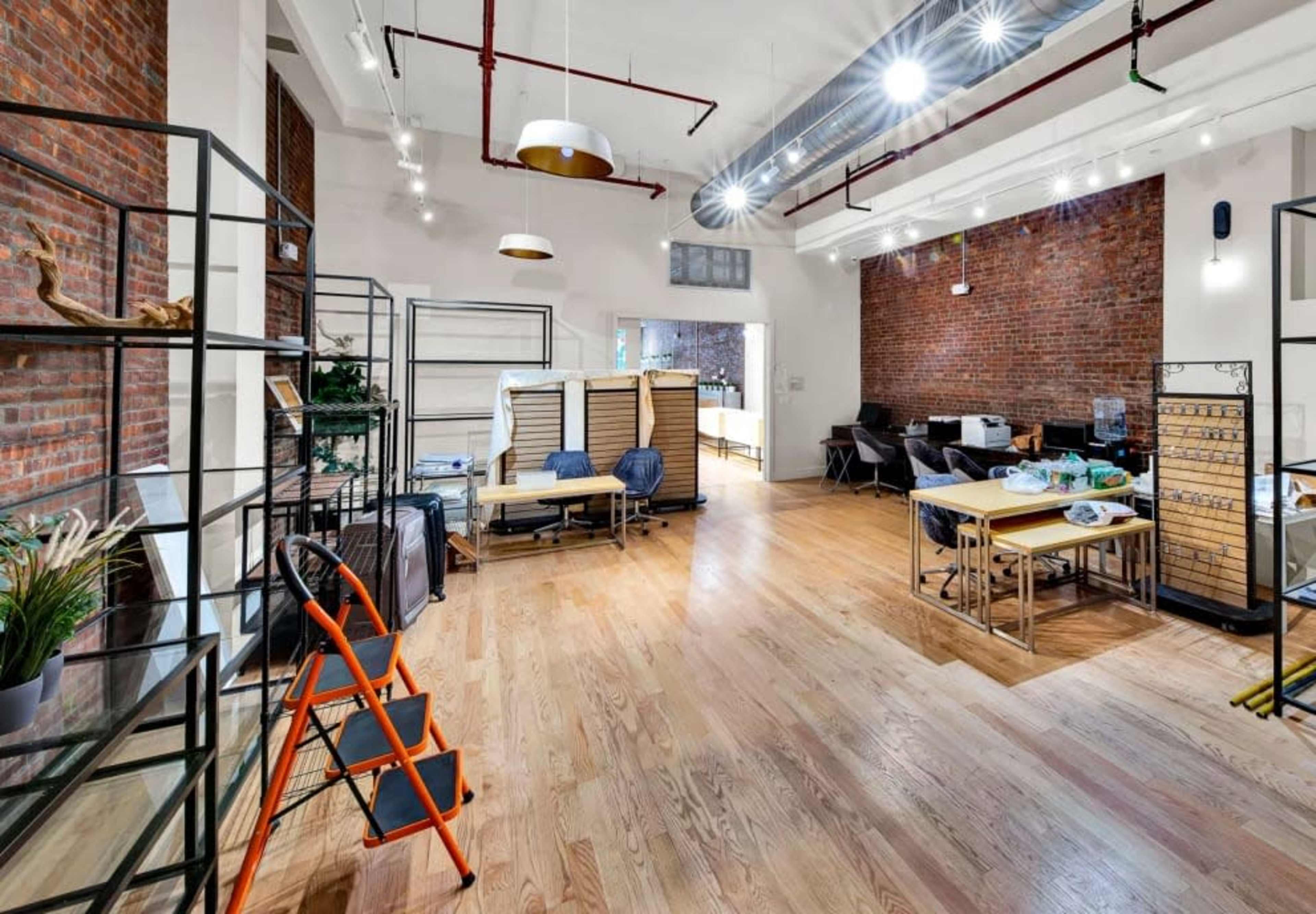 The image shows a spacious, modern interior with exposed brick walls, wooden flooring, and various furniture setups, including tables and shelving units.
