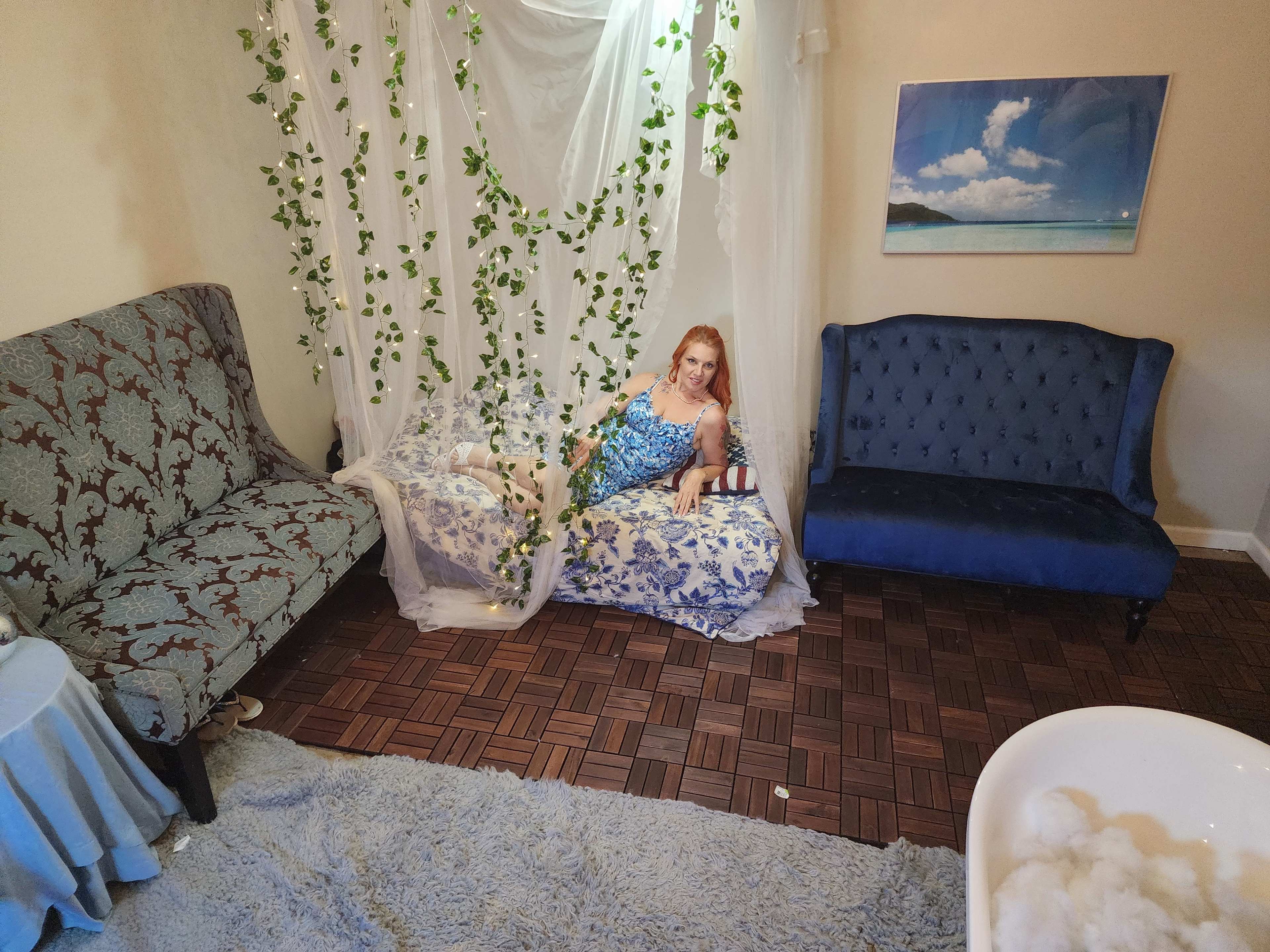 A woman in a blue dress reclines on a sofa surrounded by decorative greenery in a softly lit room.
