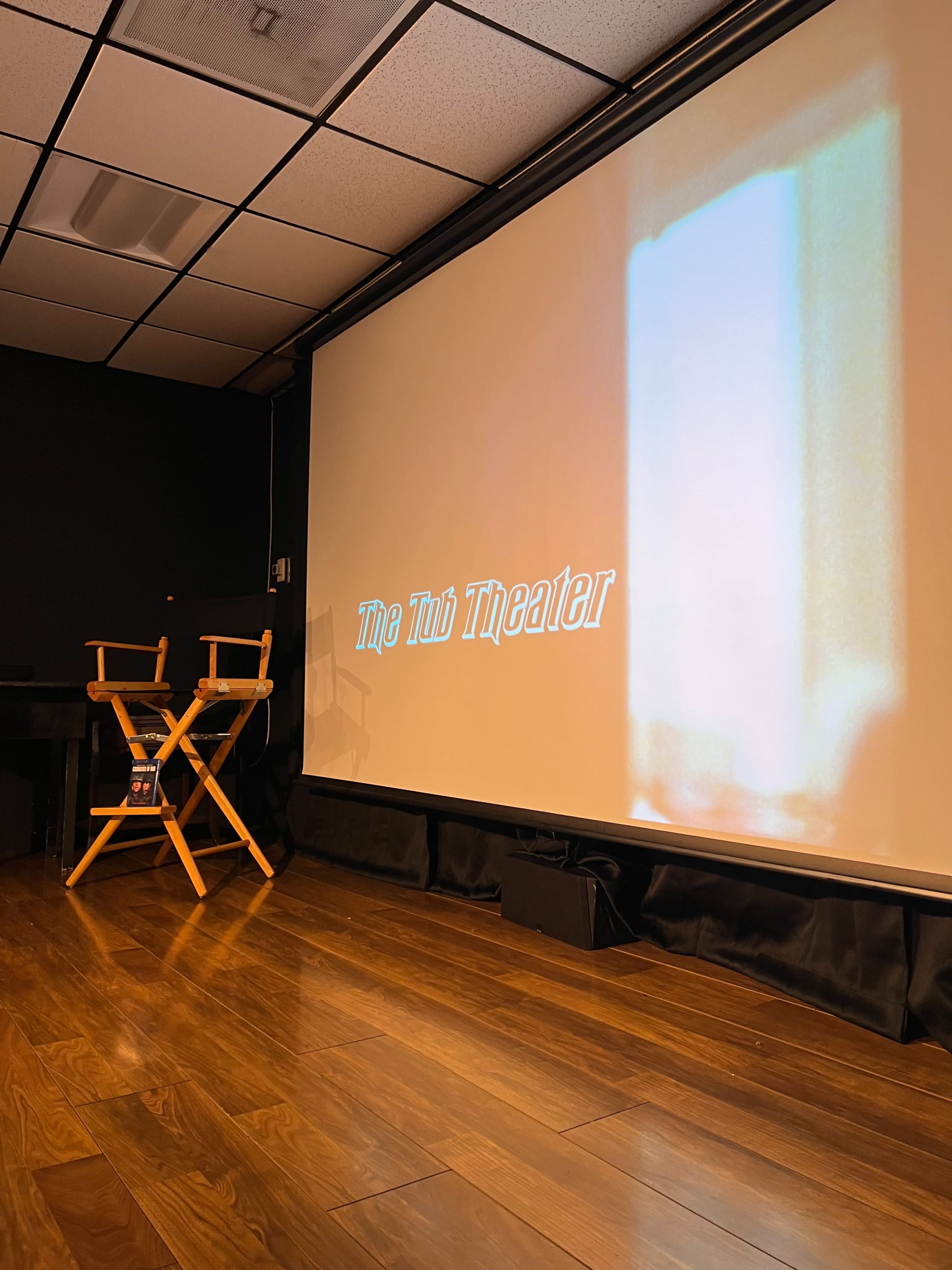 The image shows a dimly lit theater with a projection screen displaying the text "The Tub Theater" and two wooden director’s chairs in front of it.