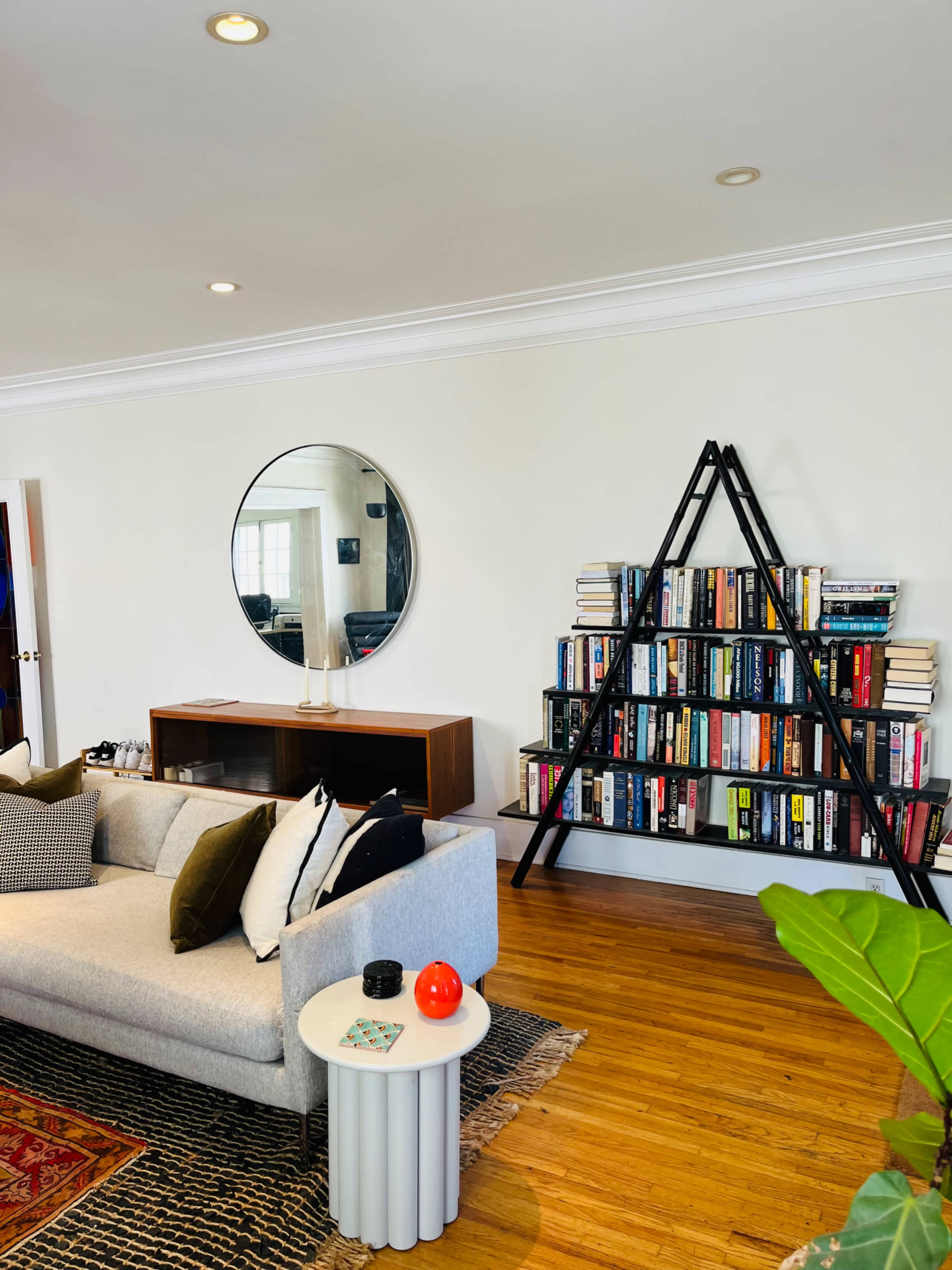 A modern living room with a gray sofa, a circular mirror on the wall, a wooden console table, and a black ladder-style bookshelf filled with books.