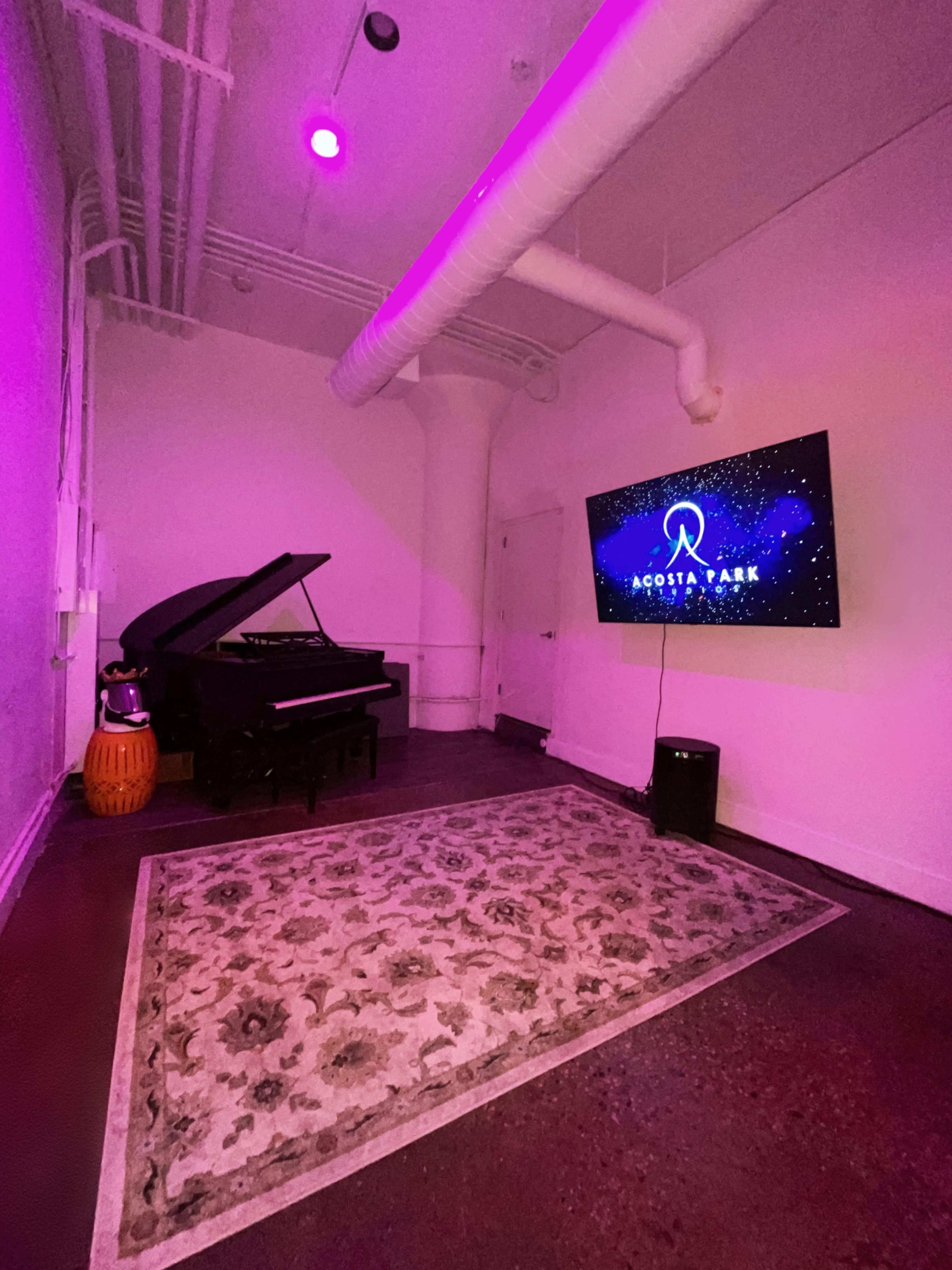 A grand piano is positioned next to a television displaying the logo for "ACOSTA PARK" in a room with purple lighting and a decorative rug on the floor.