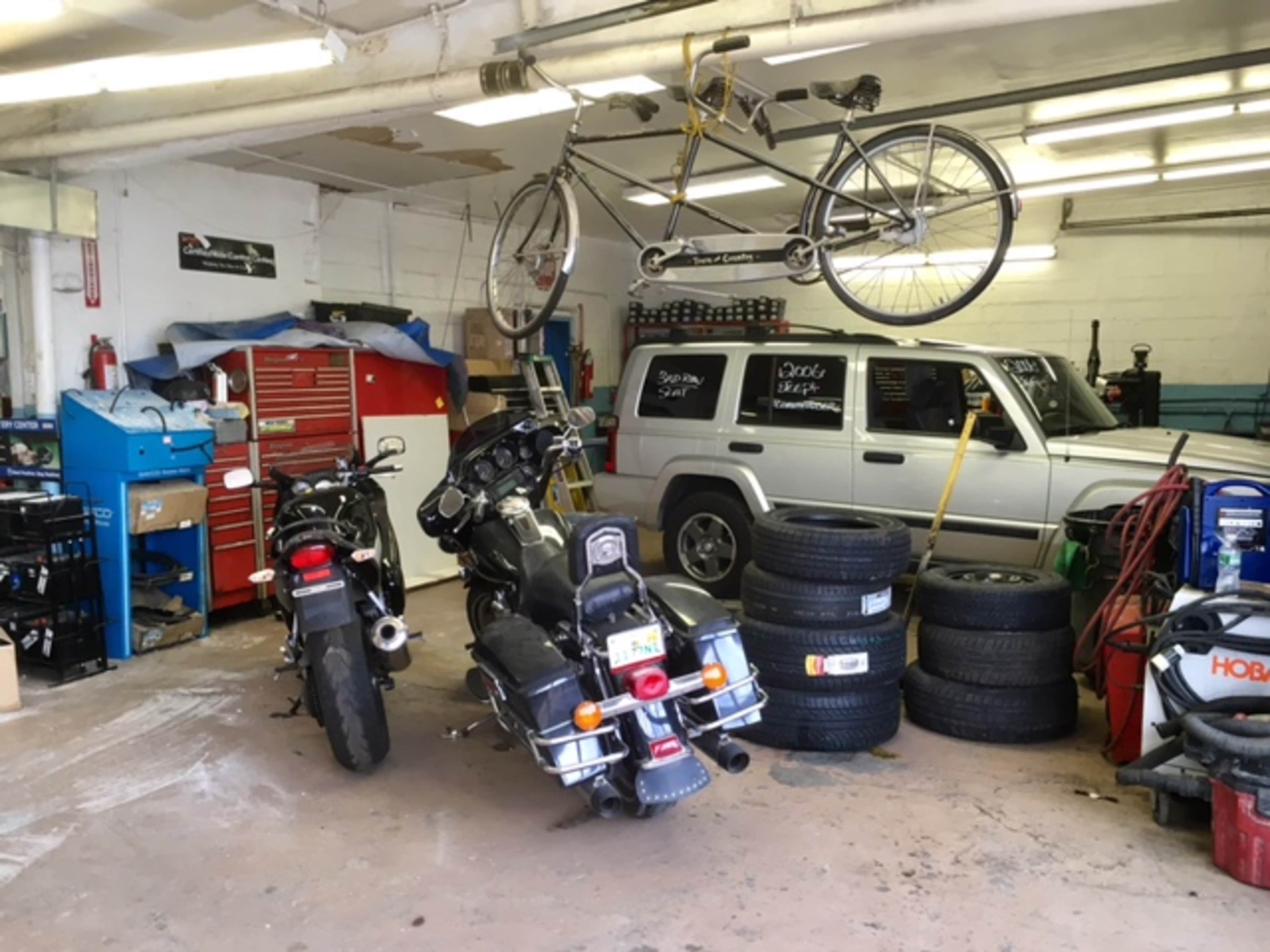 A garage with two motorcycles parked side by side, a bicycle hanging from the ceiling, and a parked SUV along with stacks of tires and various tools.