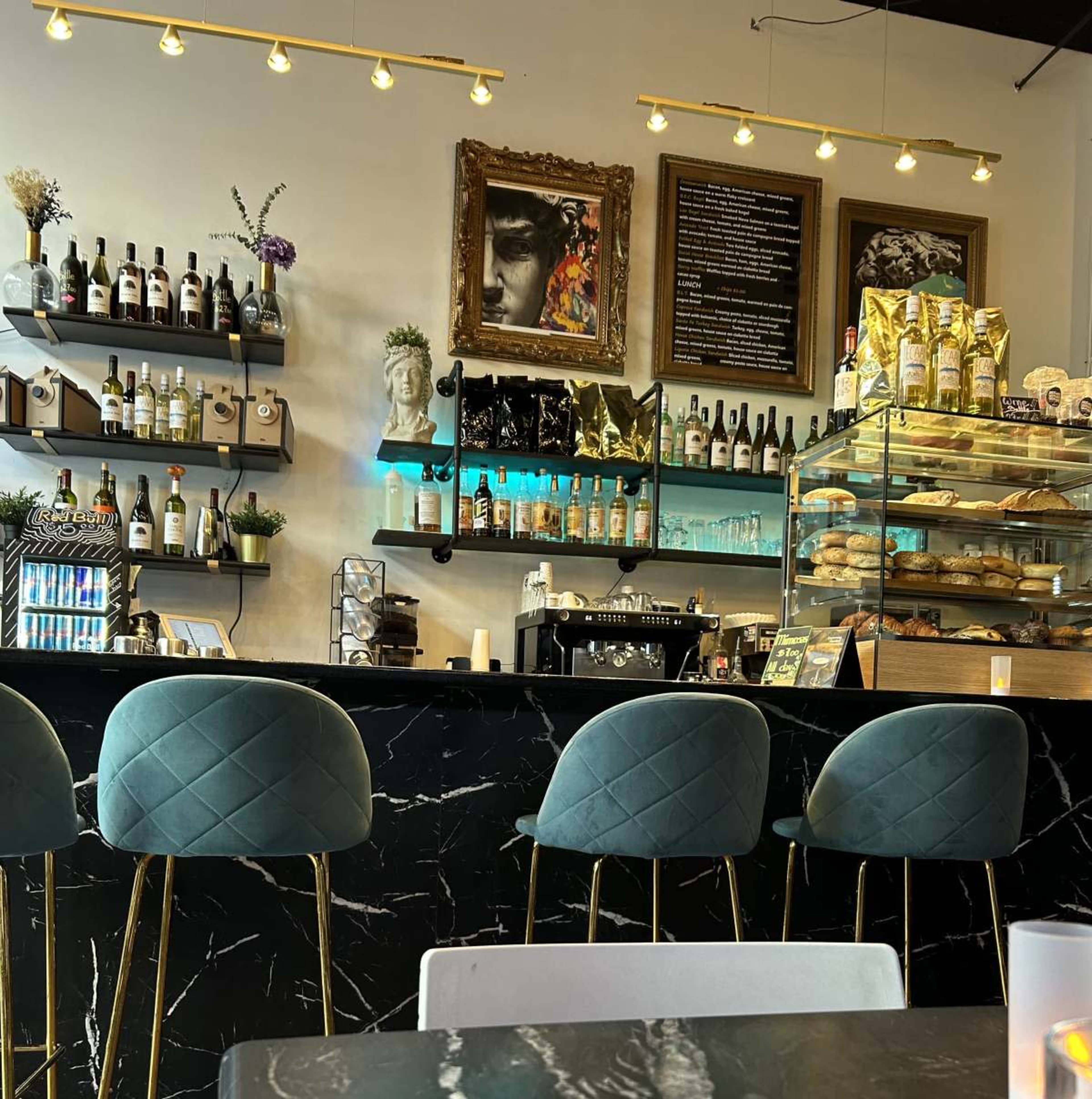 The image shows a stylish café interior with a dark marble counter, green upholstered bar stools, shelves lined with bottles, and a display case filled with pastries.