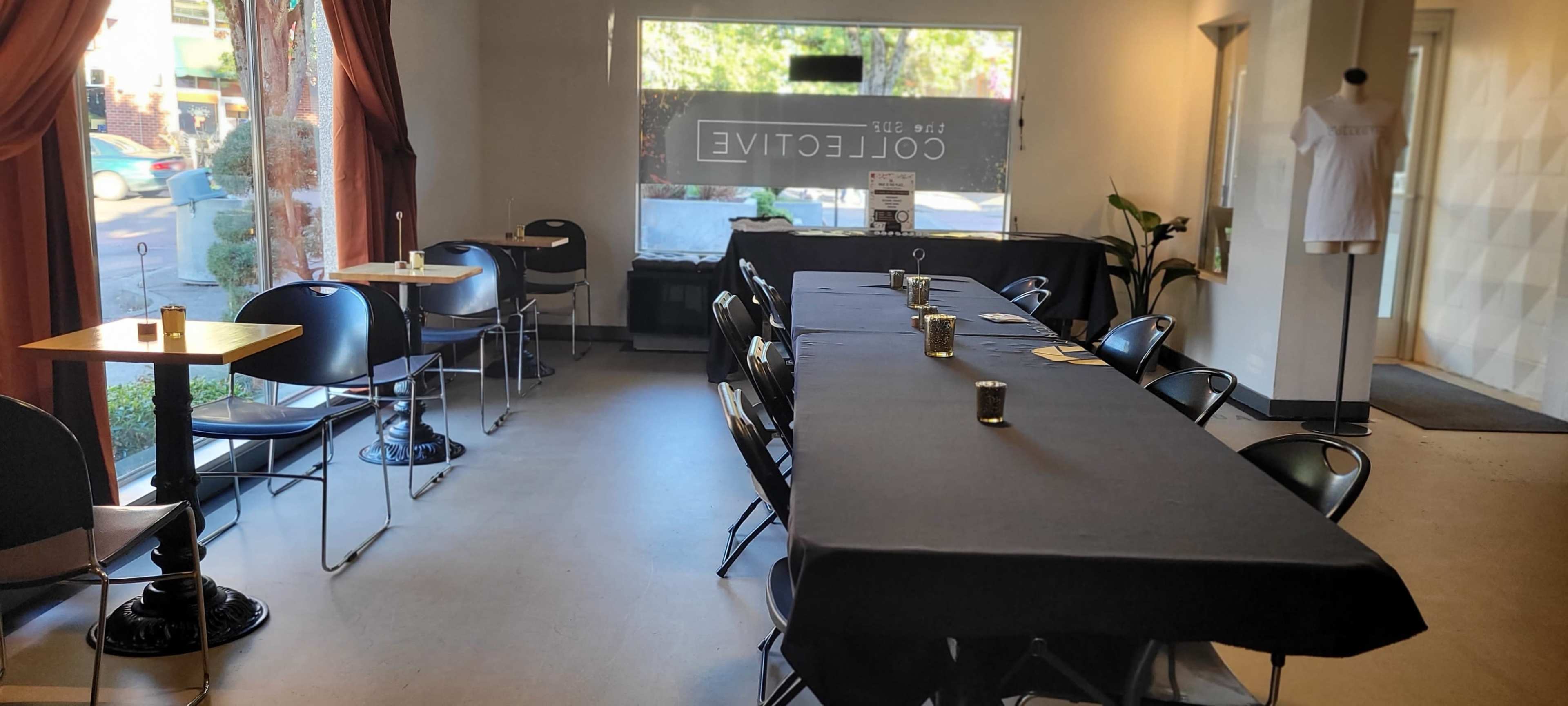 An interior of a café with a long table set up in the center, surrounded by empty black chairs, and large windows allowing natural light to enter.