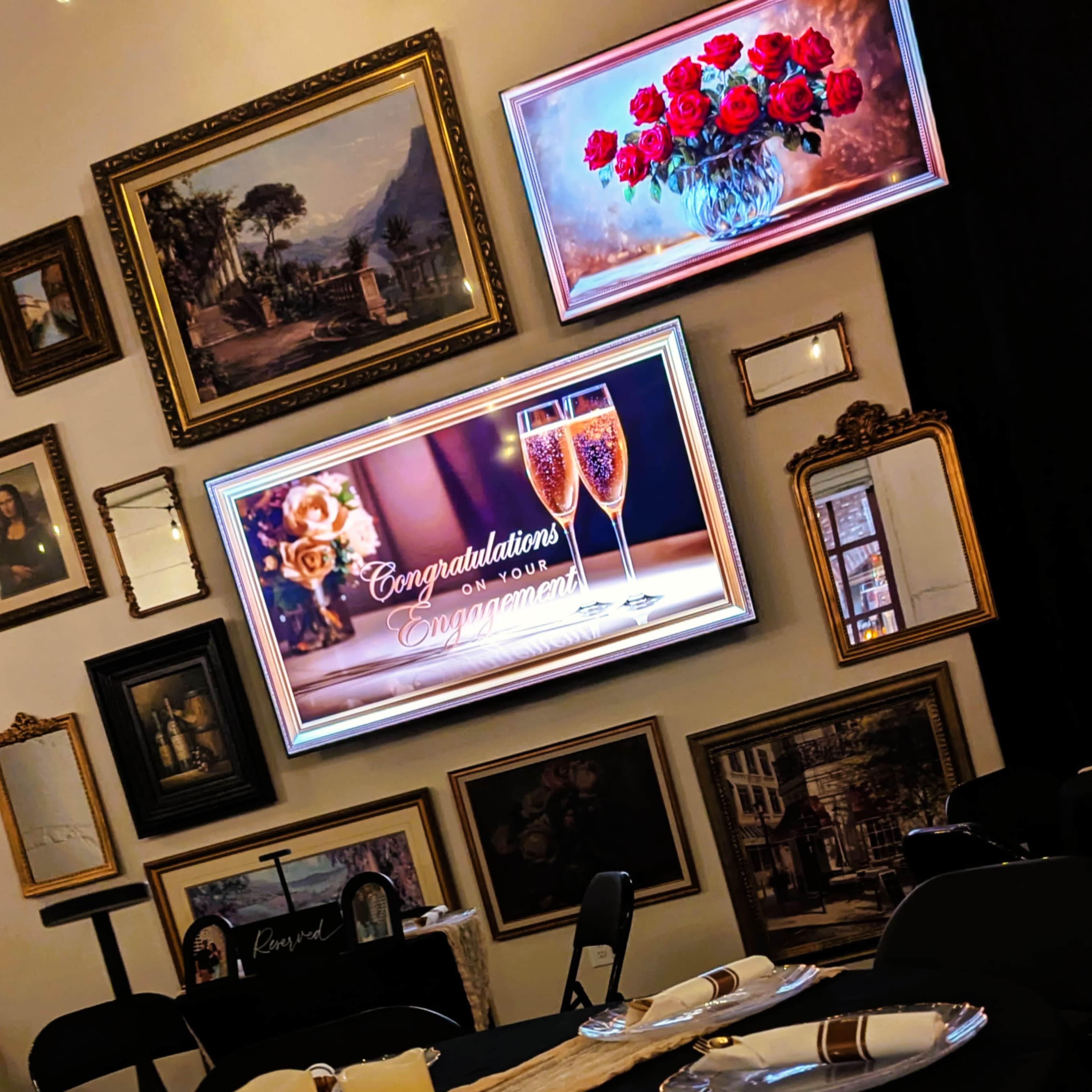 The image shows a decorated wall featuring multiple framed artworks and two digital displays, one with a bouquet of roses and a toast, and the other reading "Congratulations on your Engagement."