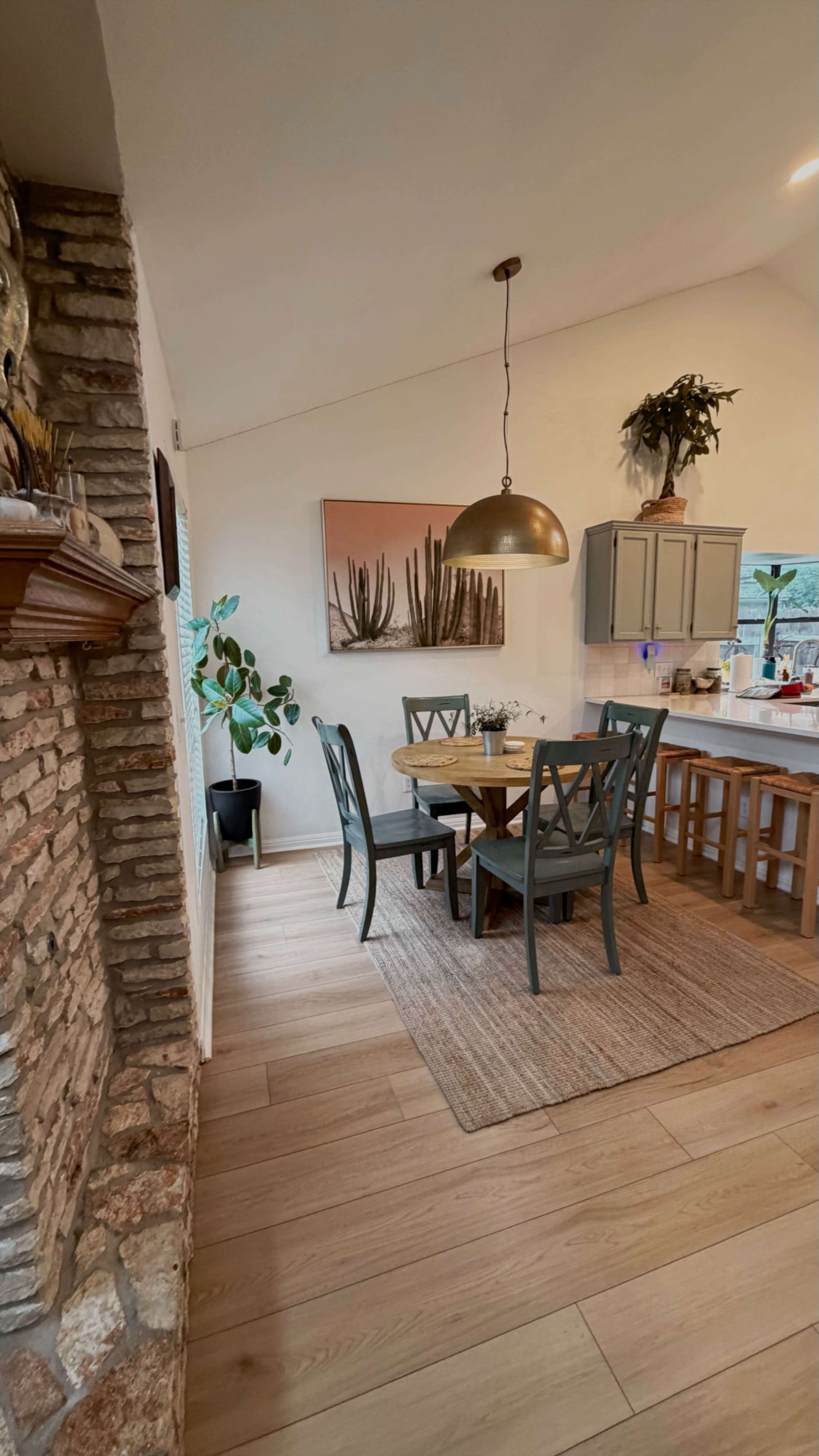 The image shows a dining area featuring a round wooden table surrounded by four gray chairs, a brick wall, and a kitchen in the background with light-colored cabinets and a large pendant light above the table.
