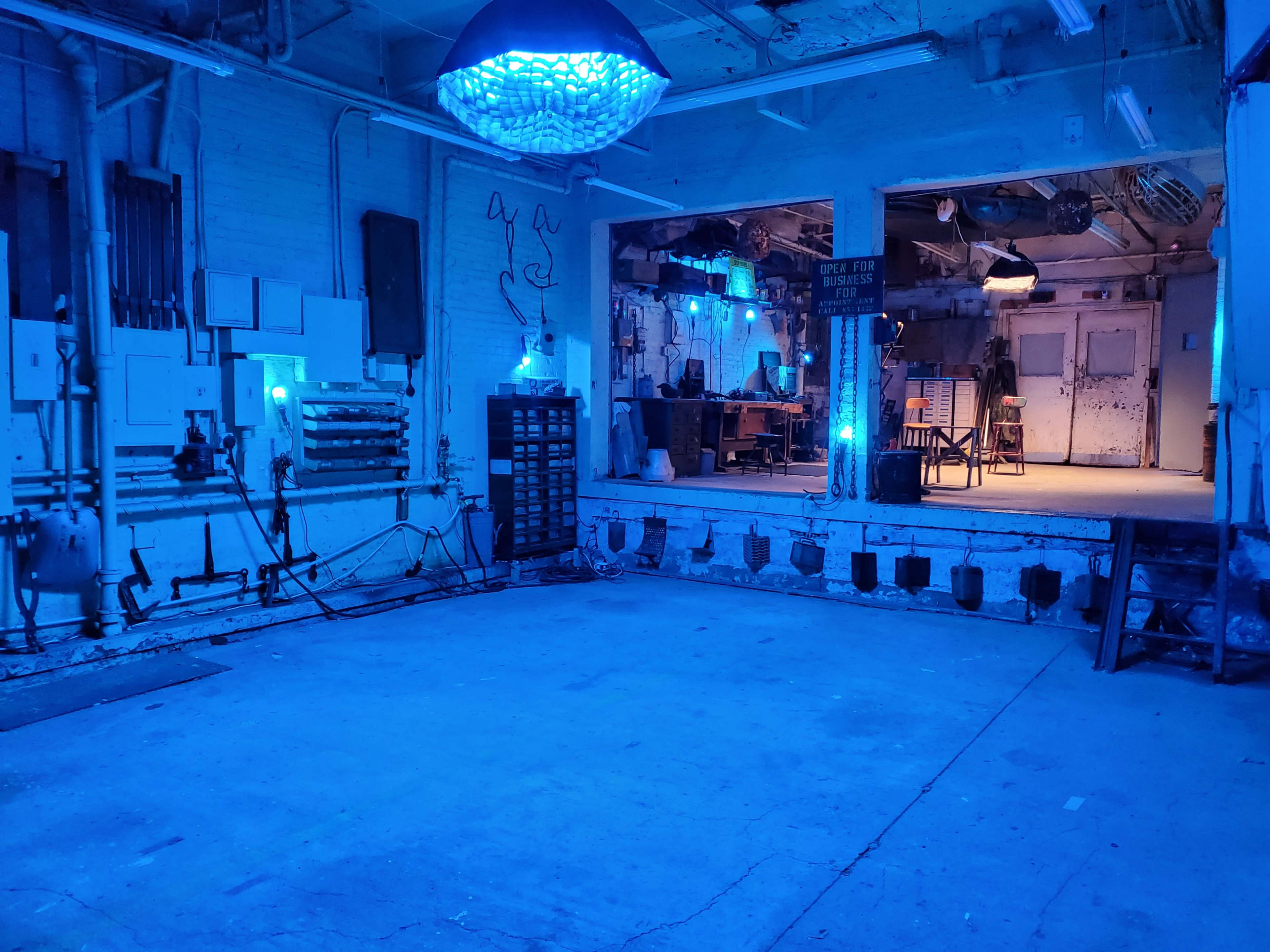 A dimly lit room with blue lighting, featuring empty concrete floors and a partially visible space with furniture in the background.