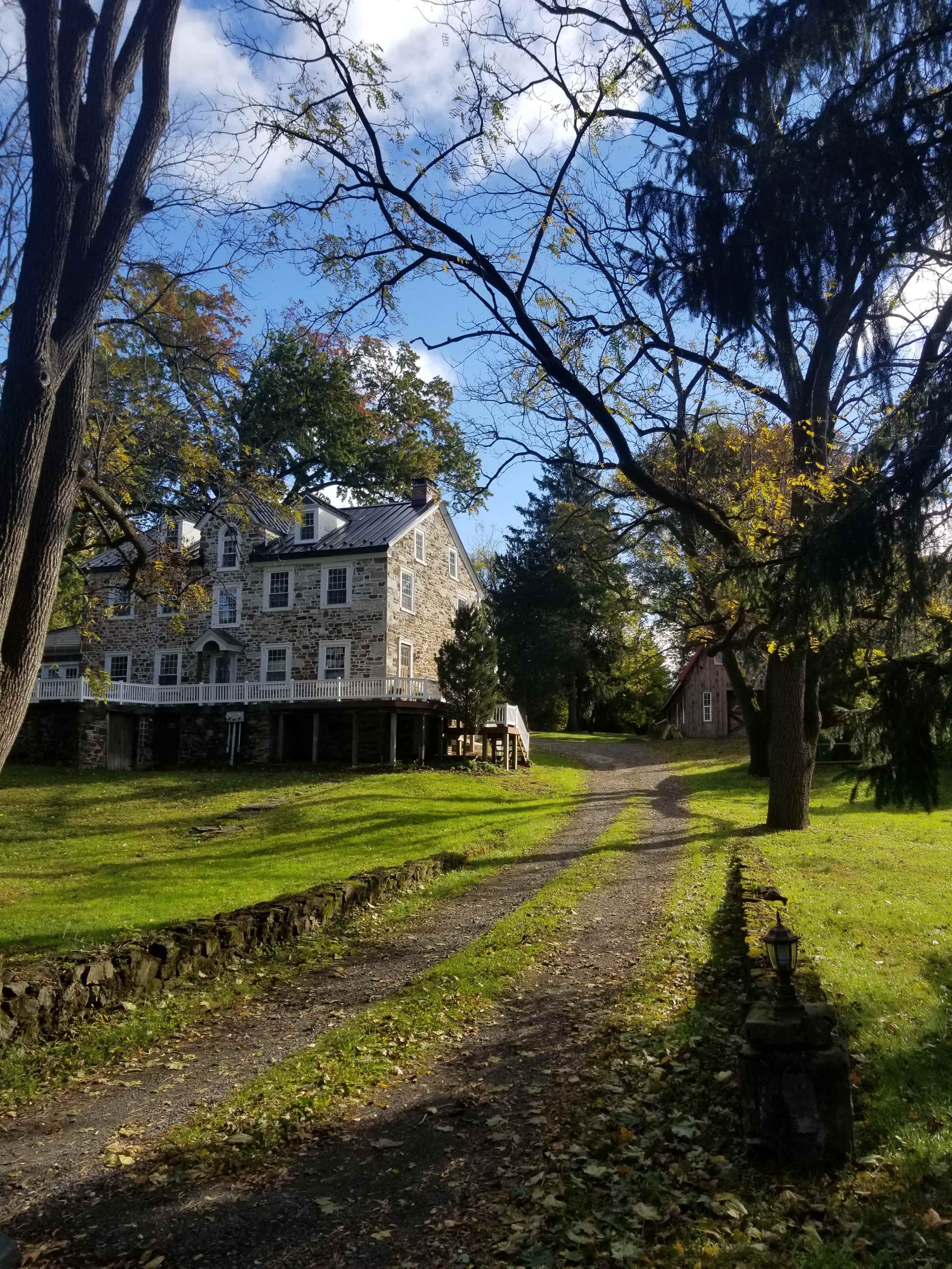 A stone house sits along a tree-lined gravel drive, with a smaller building visible in the background.