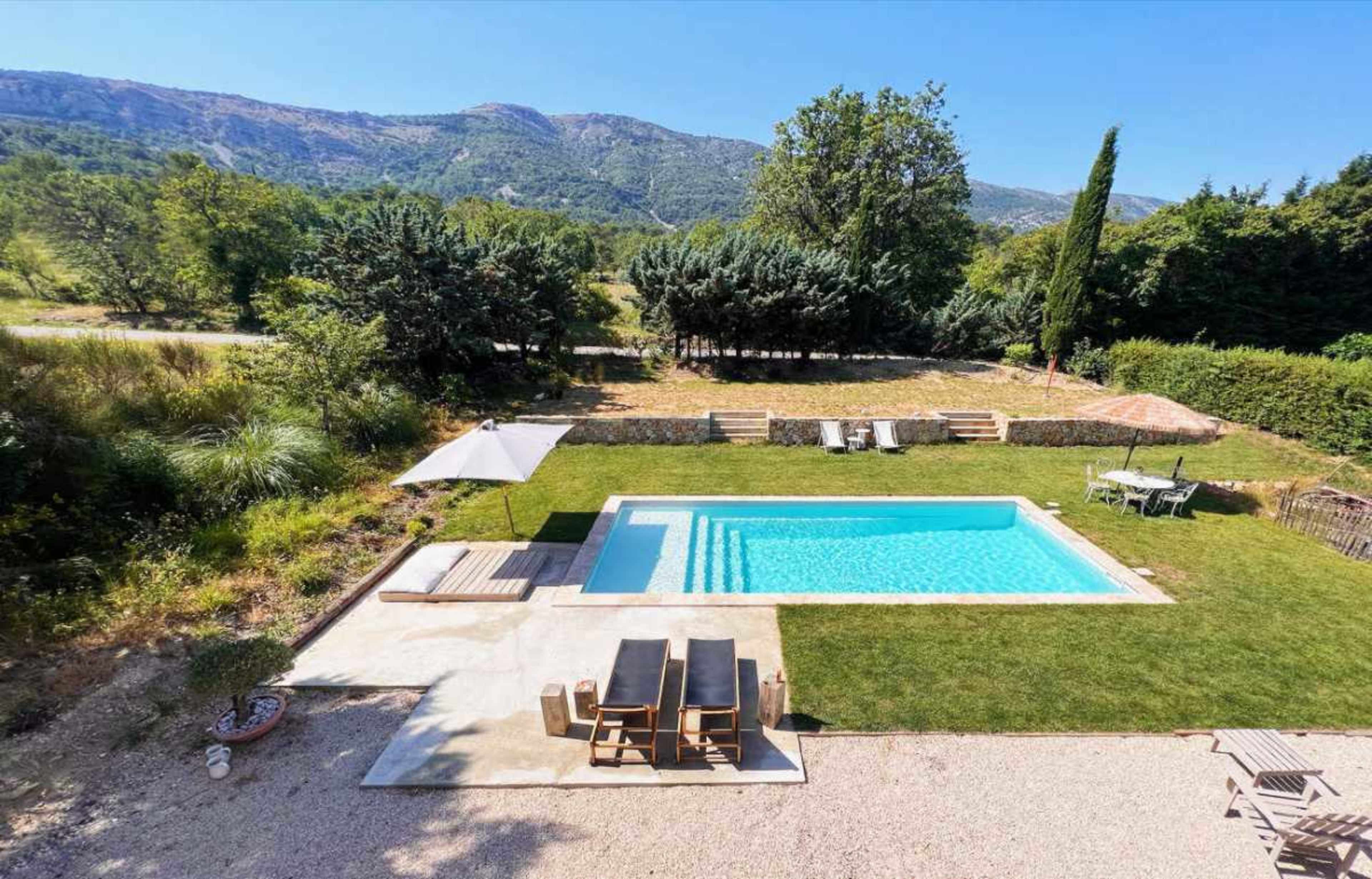 The image shows a rectangular swimming pool surrounded by a stone patio, grass, and outdoor furniture, set against a backdrop of mountains and trees.