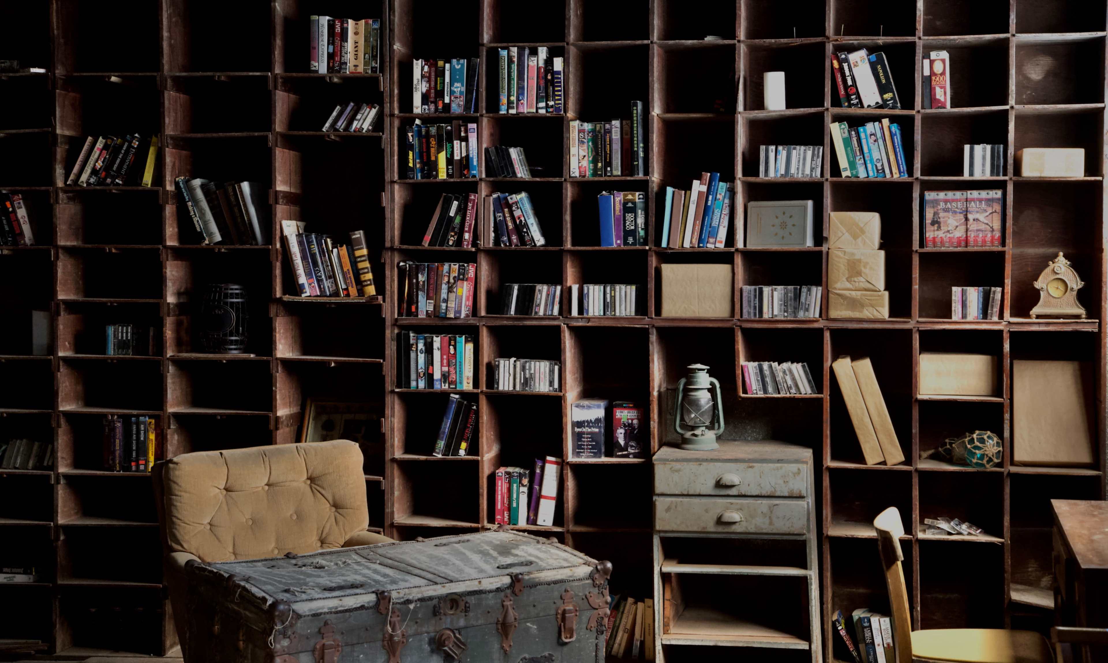 A wooden bookshelf filled with various books, CDs, and DVDs stands against a wall in a dimly lit room, with a vintage trunk and a small drawer unit in the foreground.