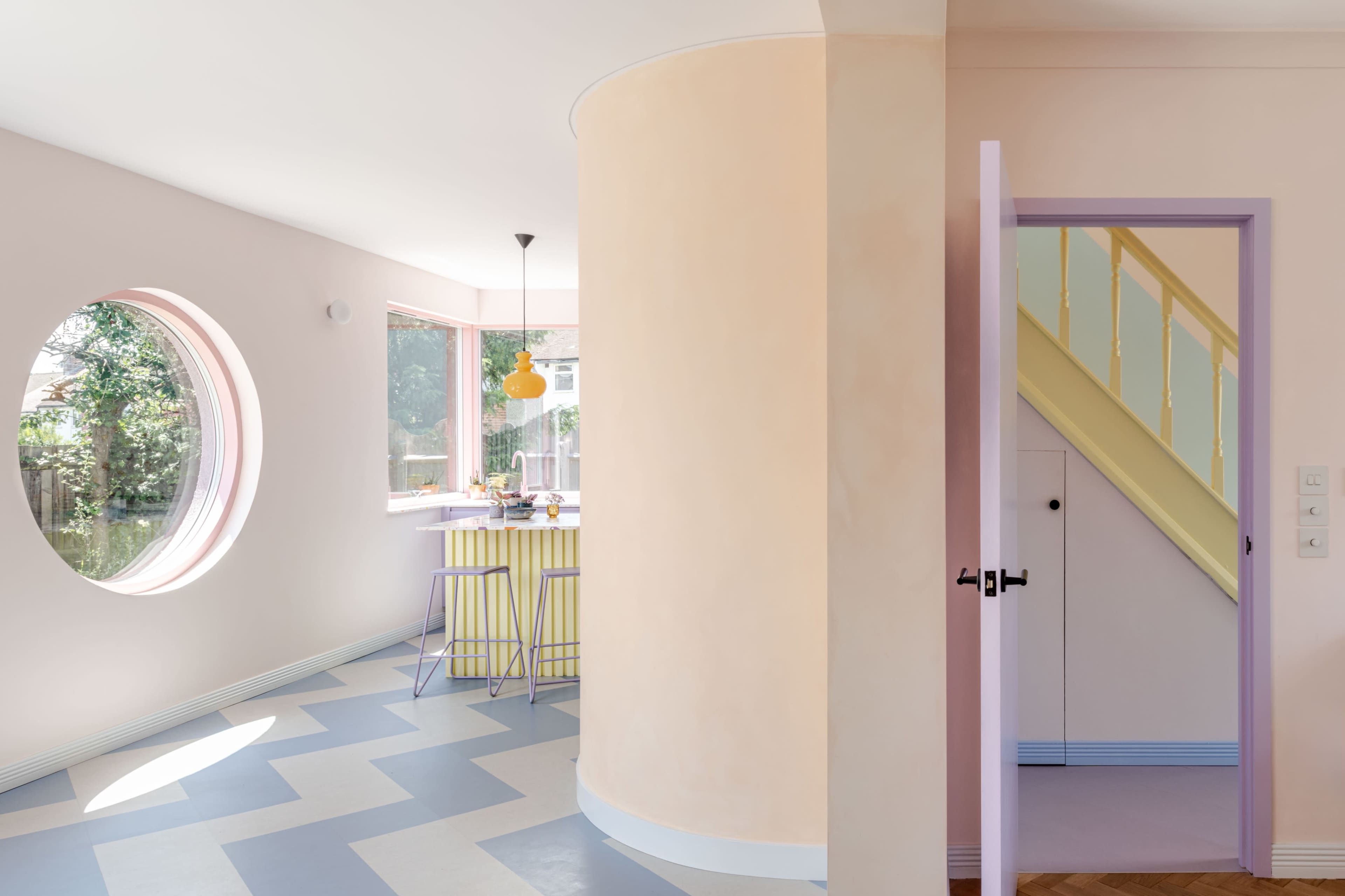 A modern interior features a circular window, pastel-colored walls, a dining area with yellow chairs, and a staircase visible through an open door.