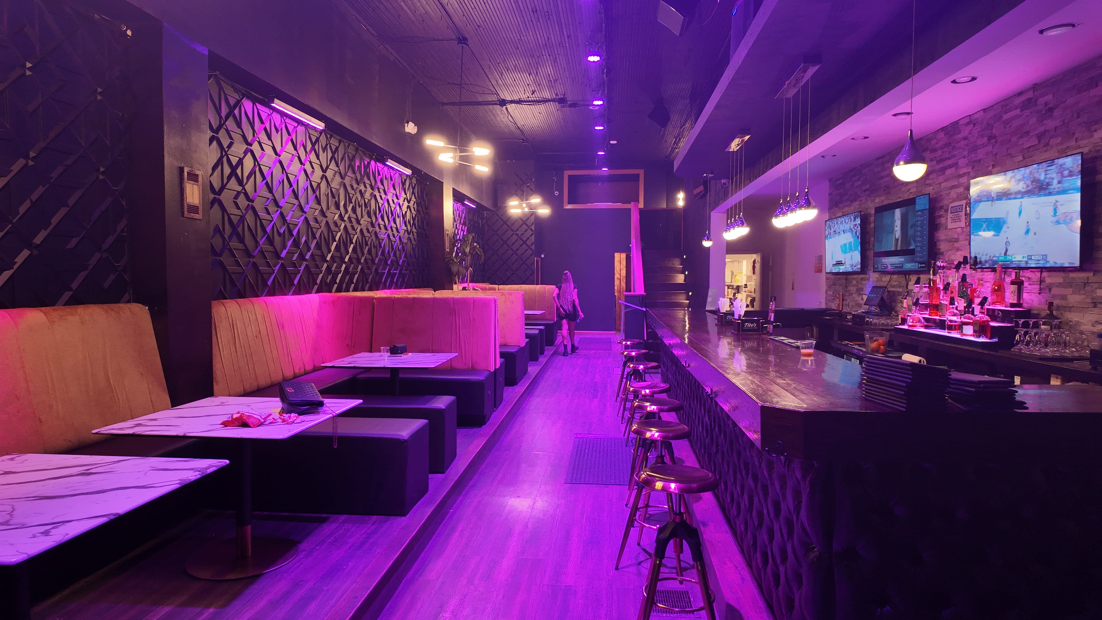 The image shows a dimly lit bar with purple lighting, featuring booth seating along one side and a long bar counter with stools on the other.