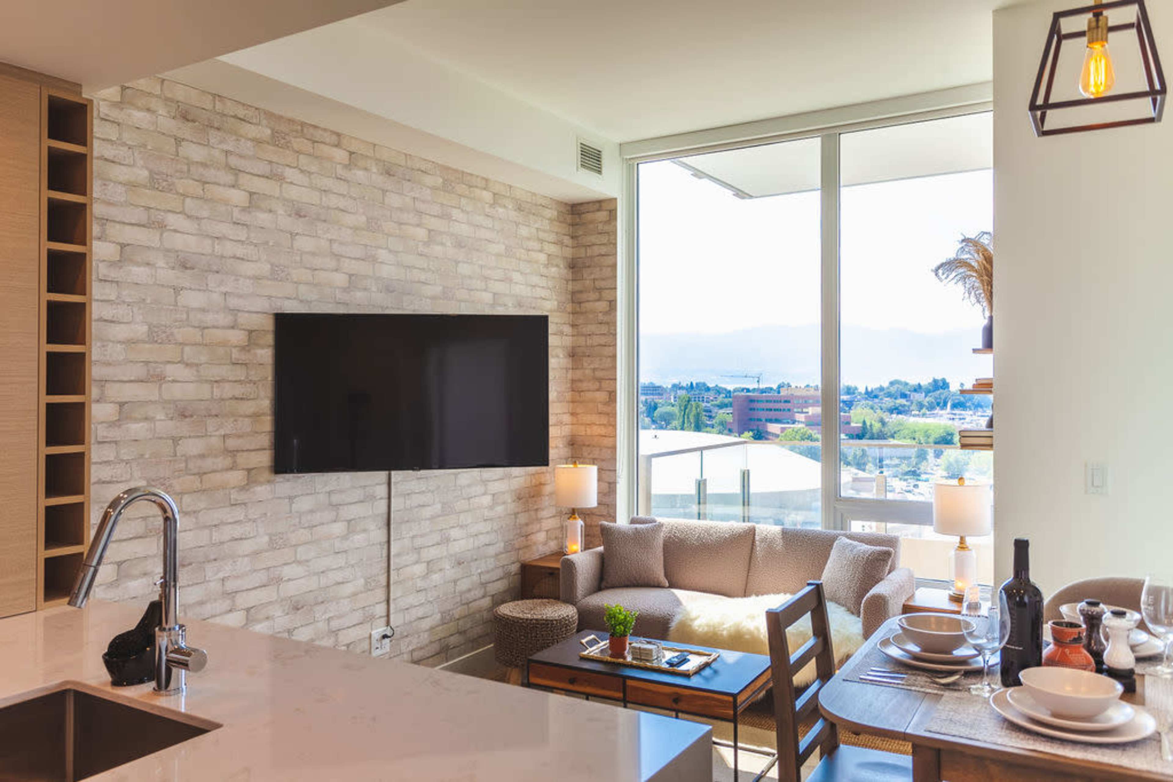 A modern living space features a kitchen area with a marble countertop, a television mounted on a brick wall, and a cozy seating arrangement by a large window with a view.