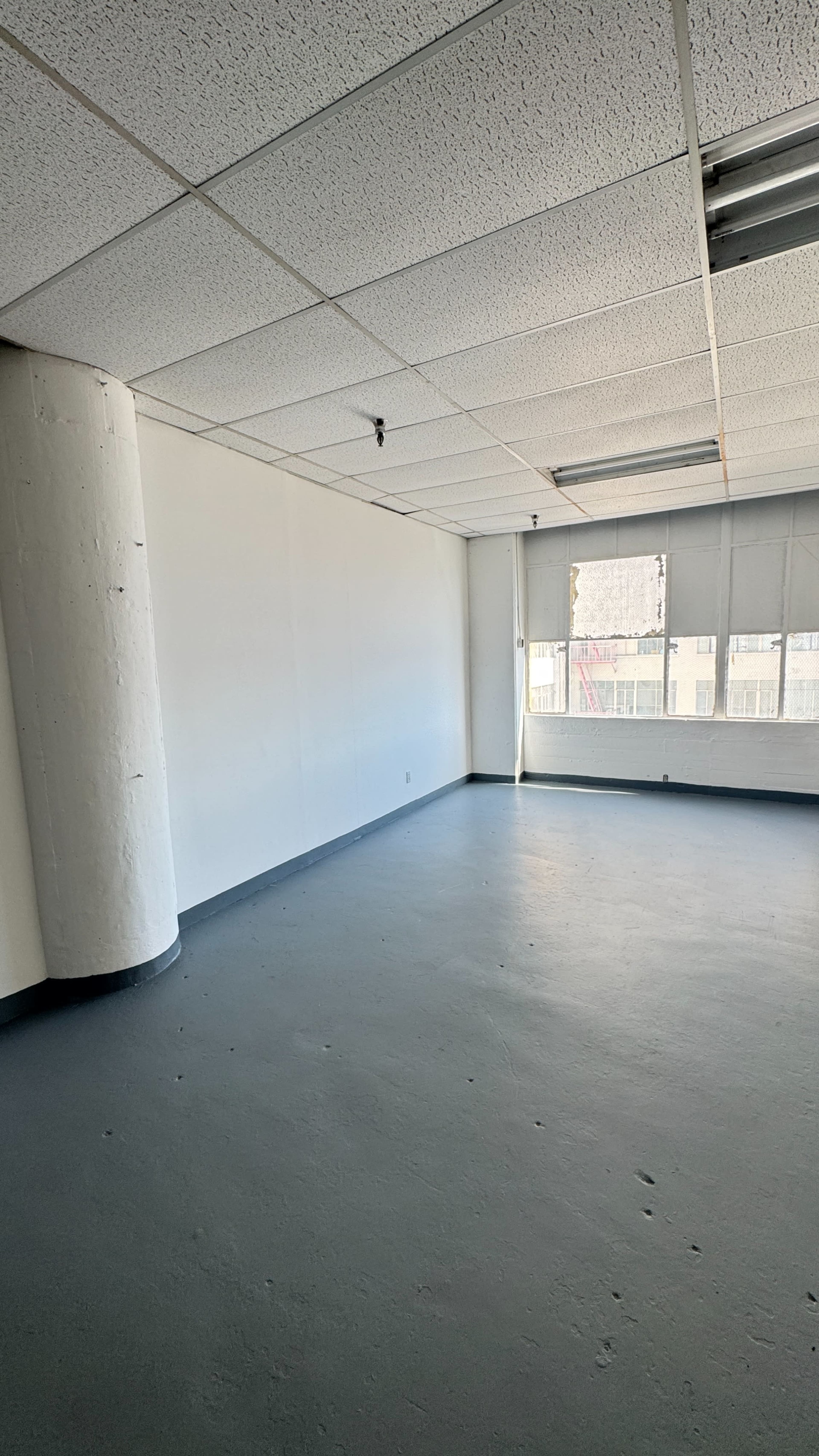 The image shows an empty room with a concrete floor, white walls, a column, and large windows allowing natural light to enter.