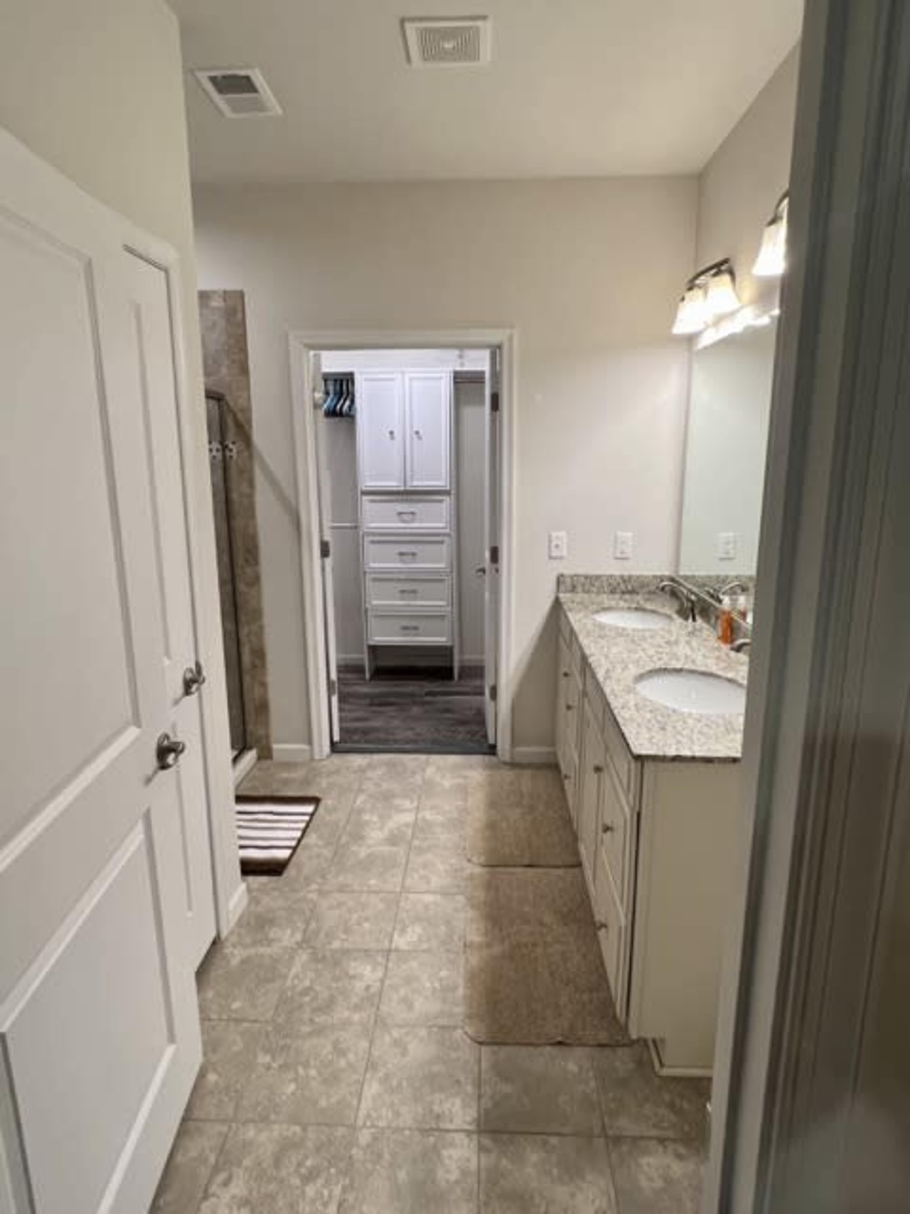 A small bathroom features dual sinks with granite countertops, light-colored walls, and a tiled floor, leading to a closet and a shower area.