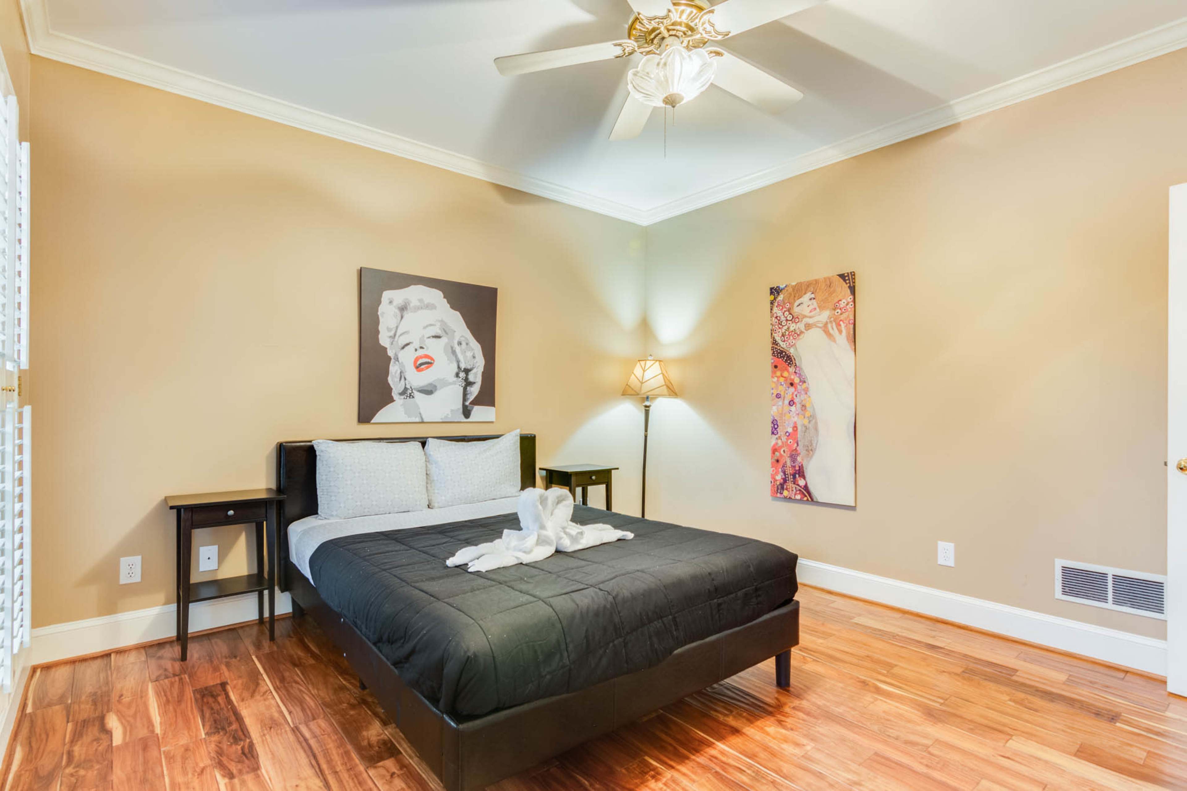 The image shows a bedroom featuring a bed with a black bedspread, two bedside tables, a ceiling fan, and framed artwork on the walls.