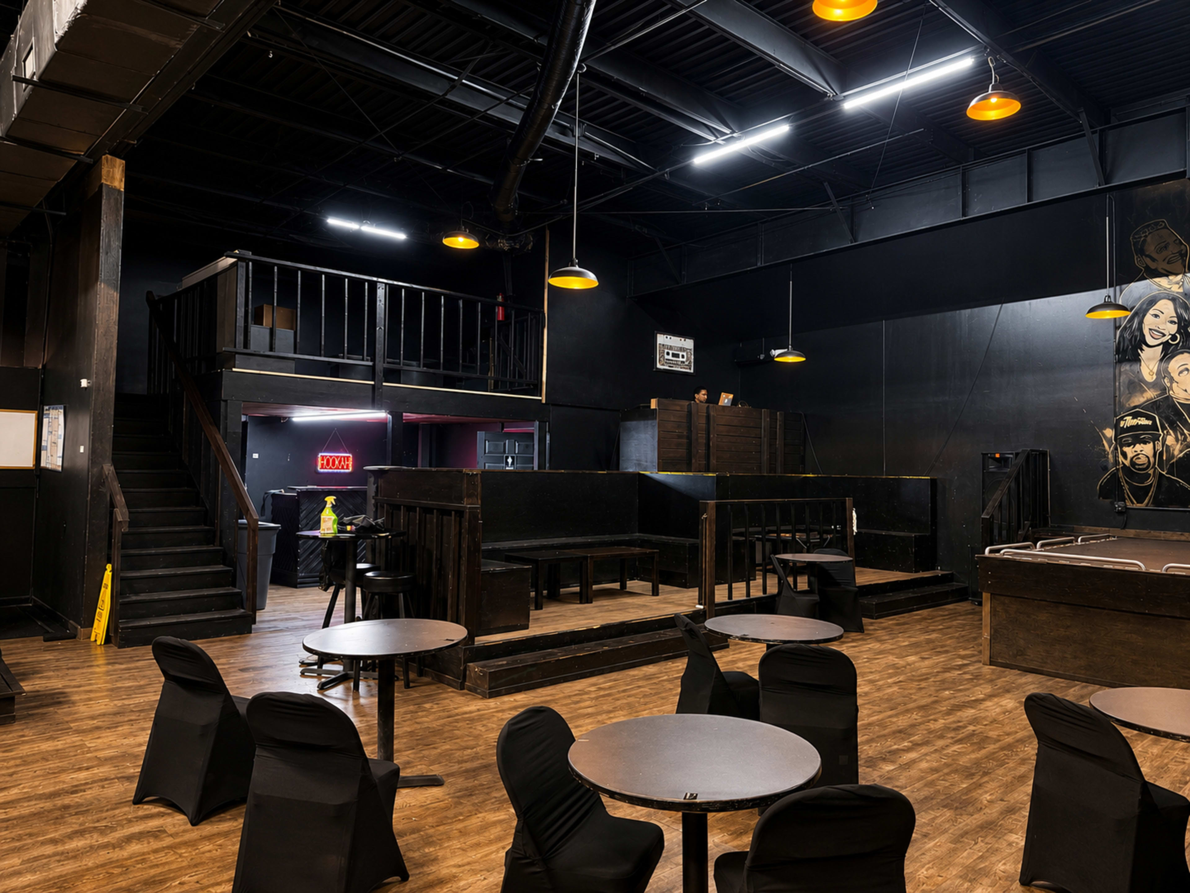 The interior of a dimly lit venue features empty tables with black chairs, a staircase leading to an upper level, and a bar area with a mural on the wall.