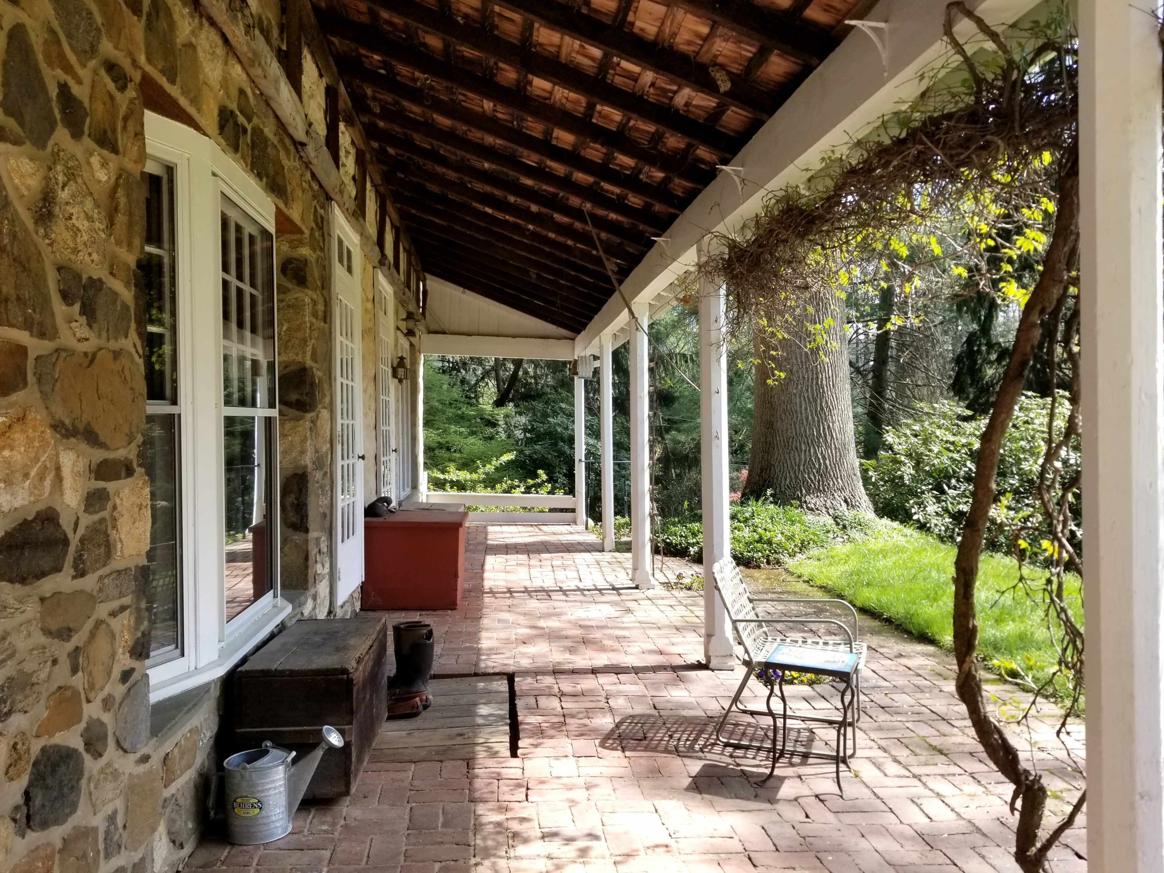 A stone porch with a tiled floor features a wooden bench and a small metal chair, surrounded by greenery.