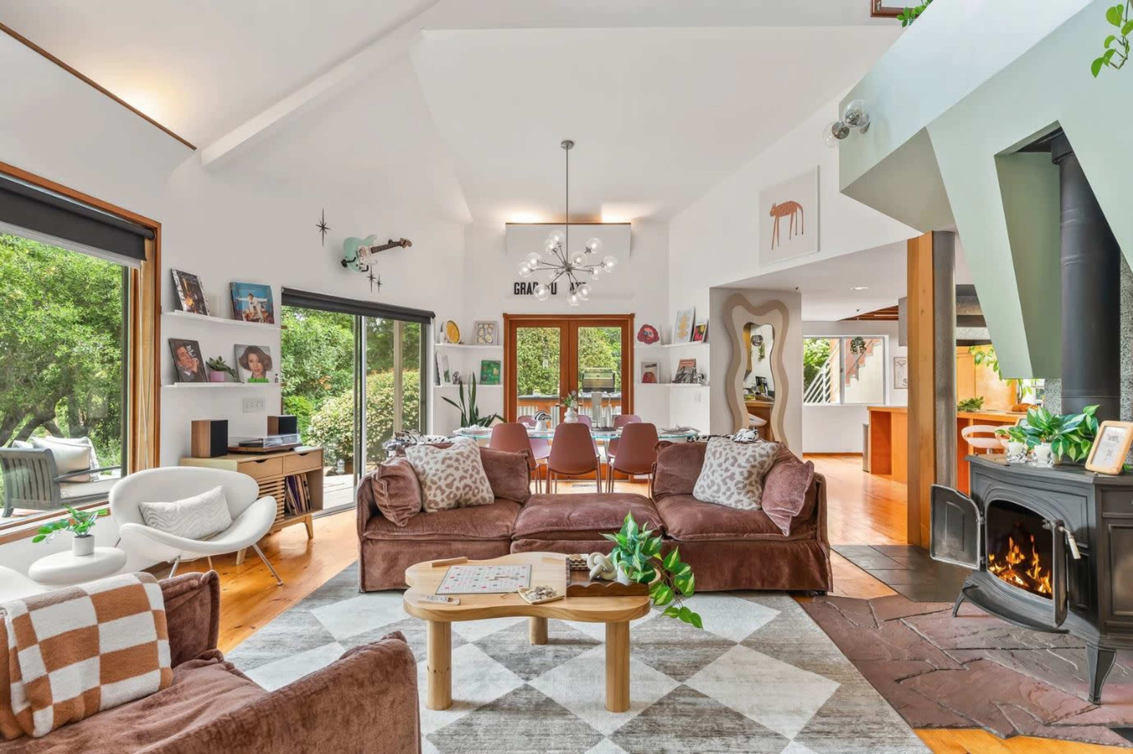 The room features a spacious living area with brown sofas, a patterned rug, and large windows overlooking greenery, alongside a dining area and a modern kitchen.