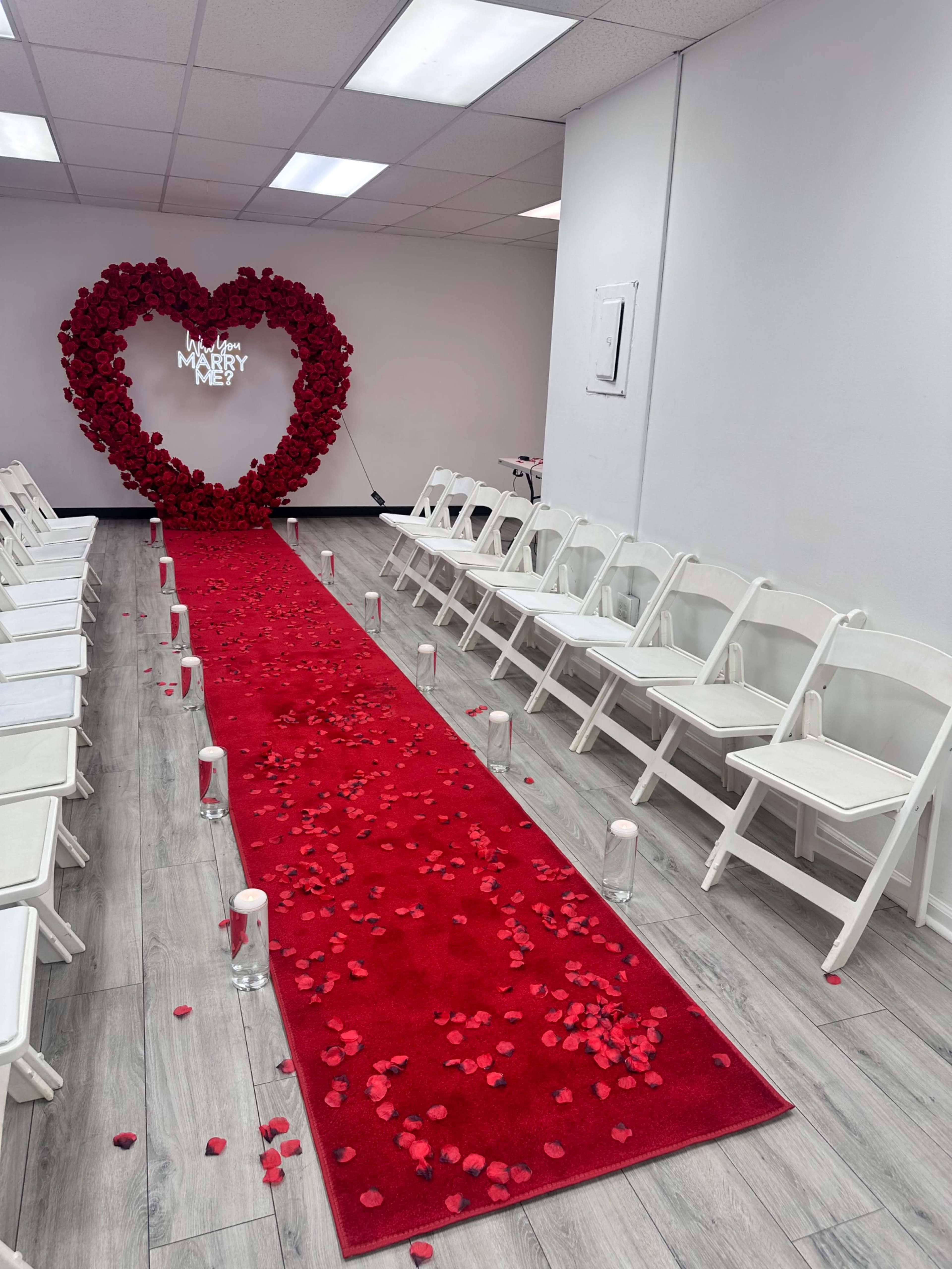 The image shows a decorated wedding ceremony area featuring a heart-shaped arrangement of roses at one end of a red carpet, with rows of white chairs on either side.