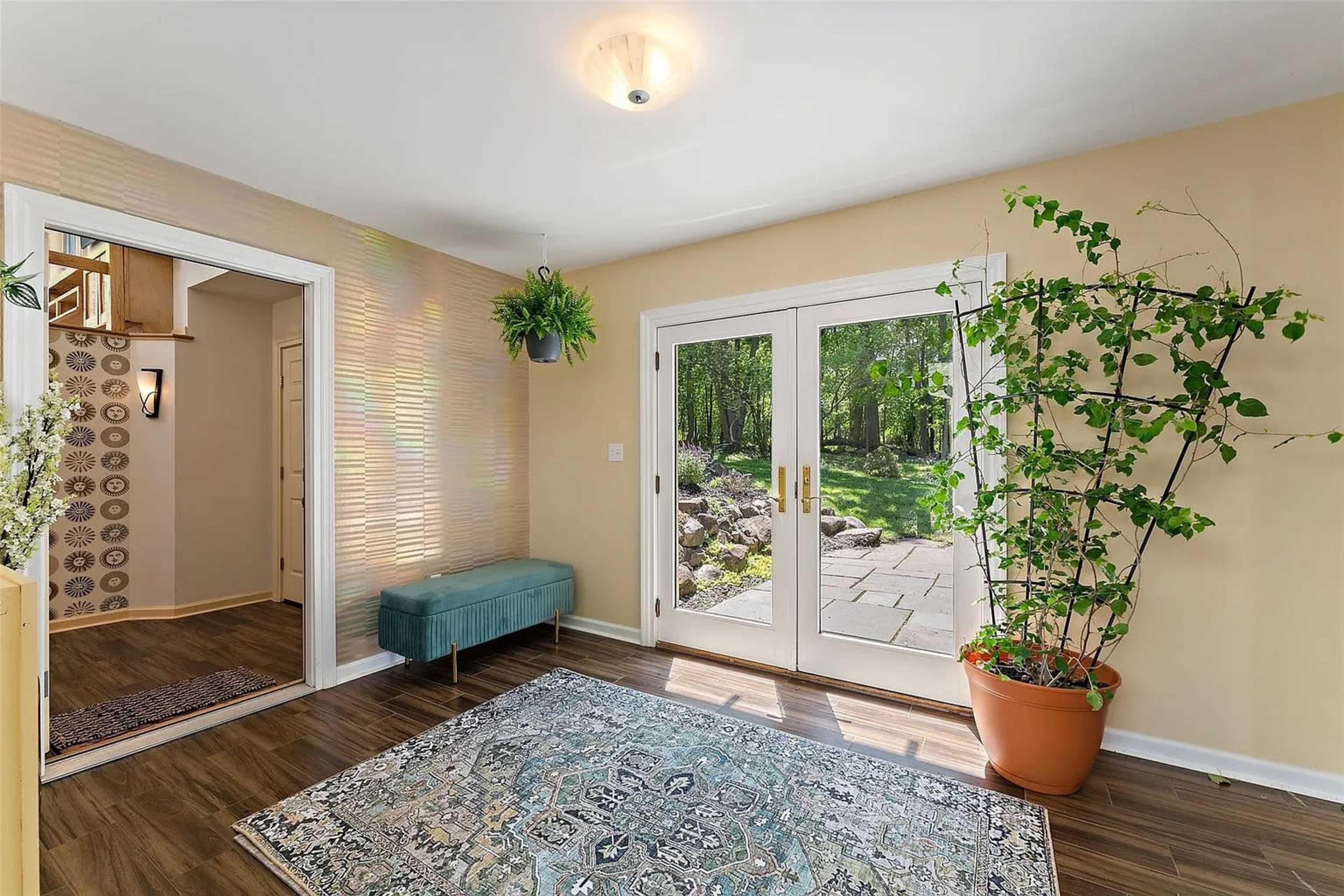 A well-lit entryway features a blue bench, a large potted plant, and double doors leading to a garden area.