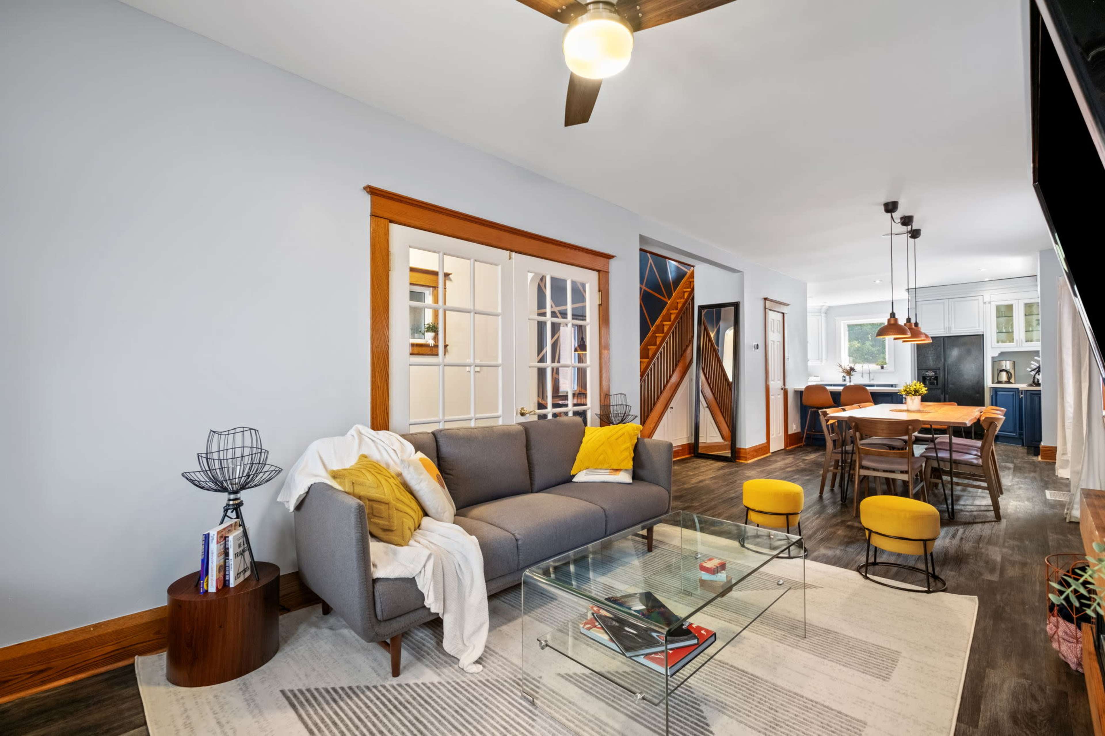 The image shows a living room featuring a gray sofa, a glass coffee table, and a dining area in the background with a wooden table and yellow chairs.