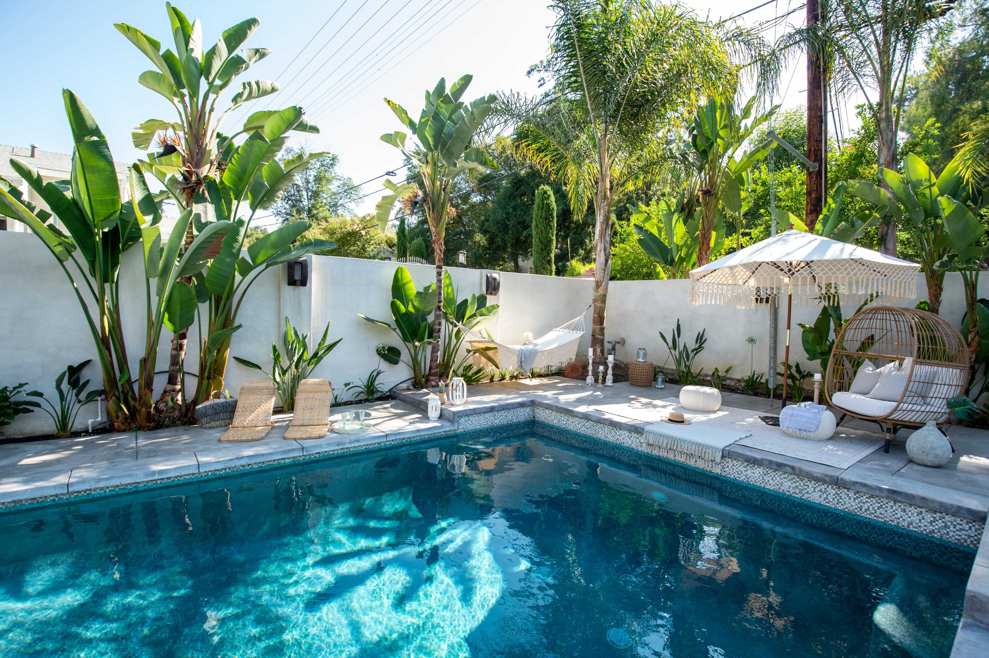 A tranquil backyard featuring a swimming pool surrounded by tropical plants, lounge chairs, and a shaded seating area with an umbrella.