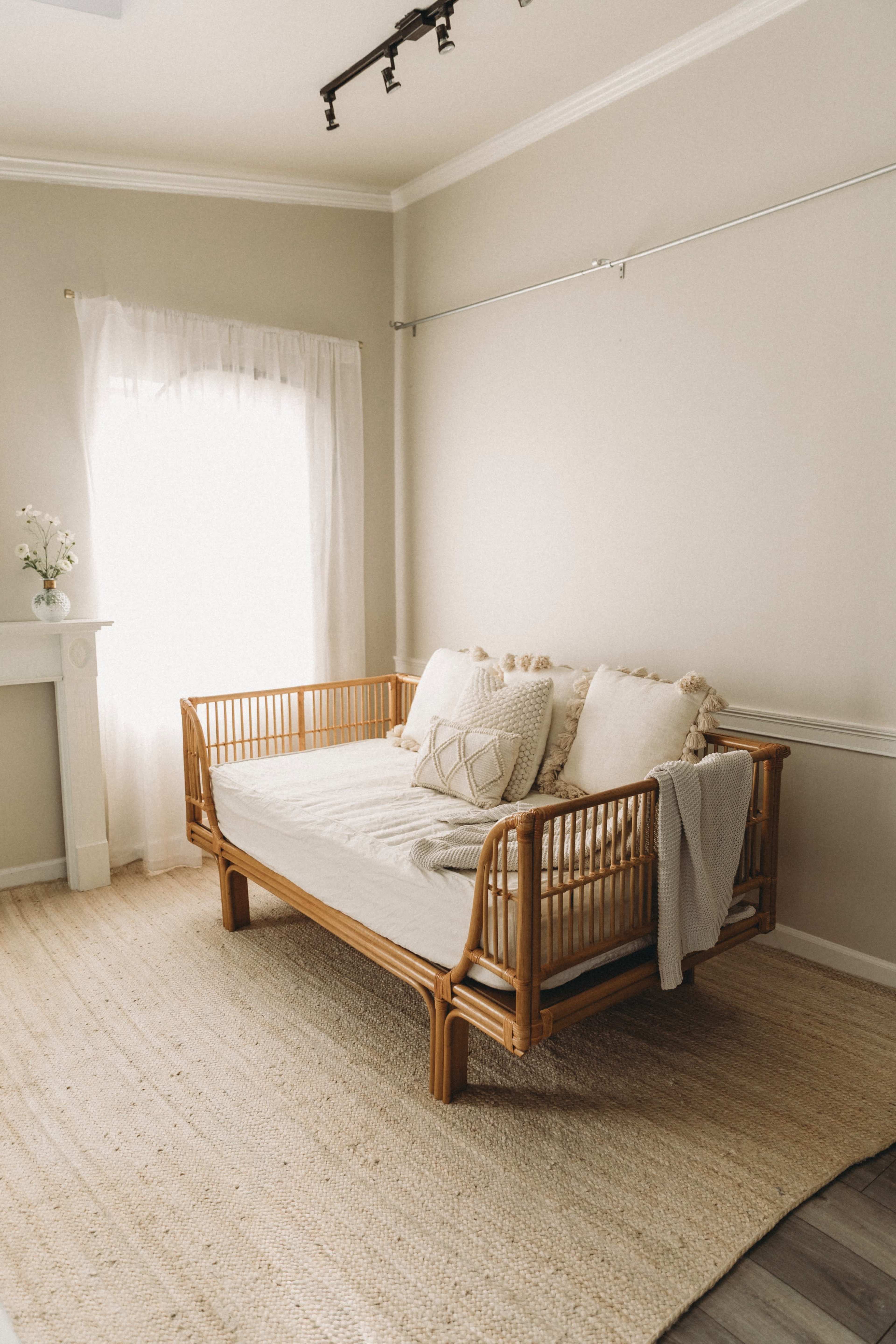 The image shows a minimalist room featuring a rattan daybed, adorned with cushions and a blanket, positioned near a large window with sheer curtains and a textured area rug on the floor.
