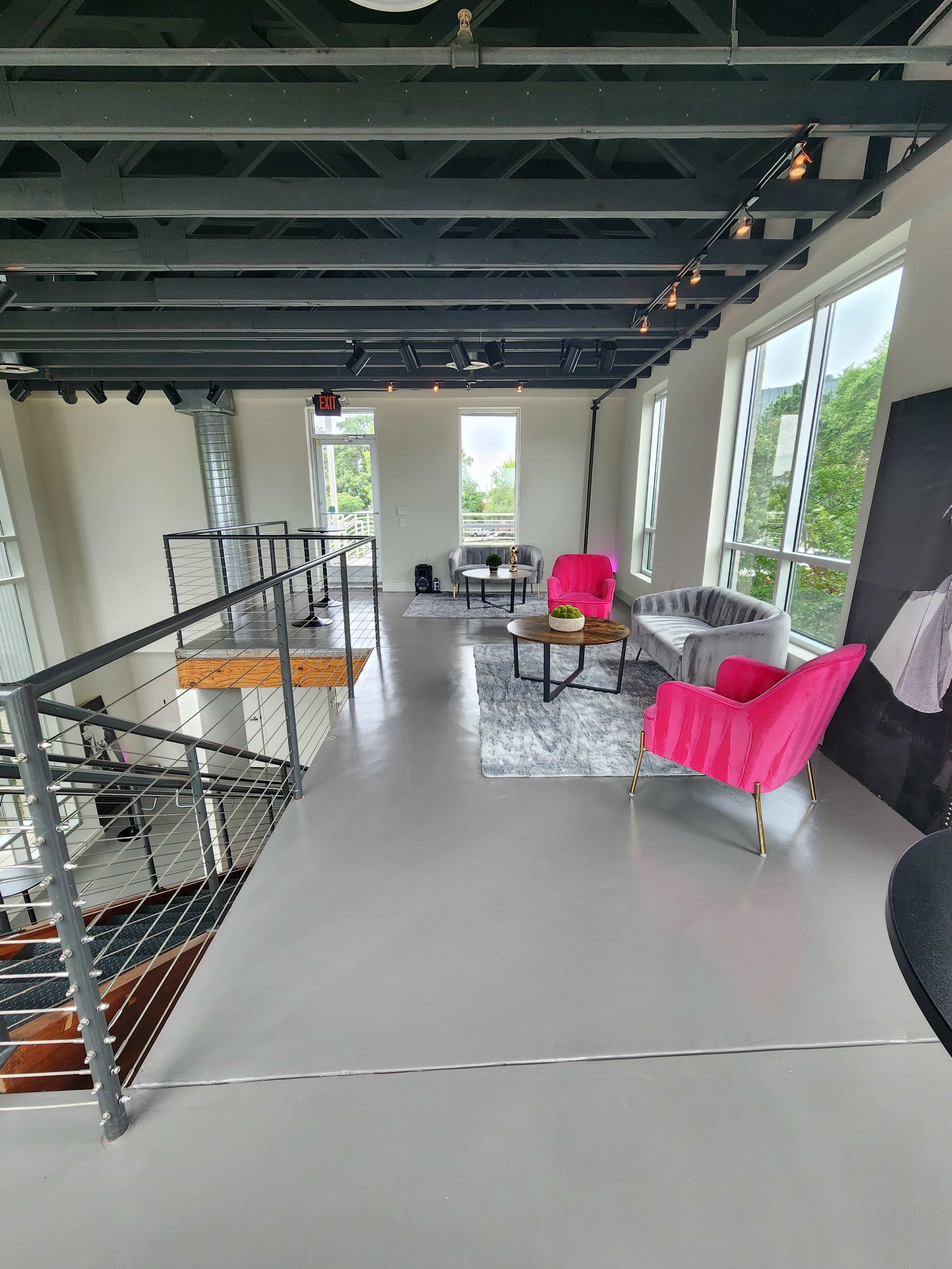 A modern, well-lit interior space featuring pink and gray seating, large windows, and a staircase.