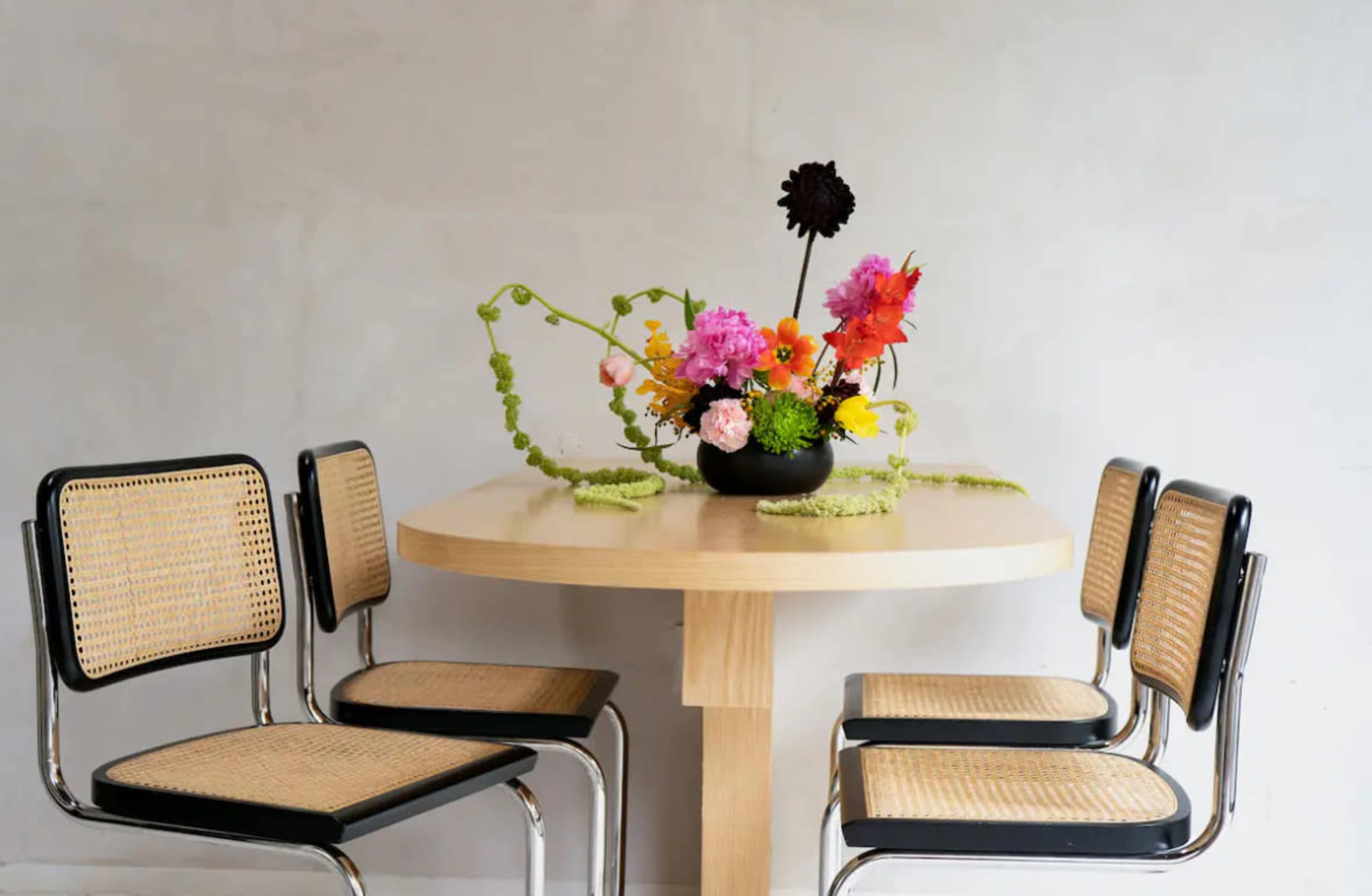 A round wooden table with a vase of colorful flowers is surrounded by four black and rattan chairs.