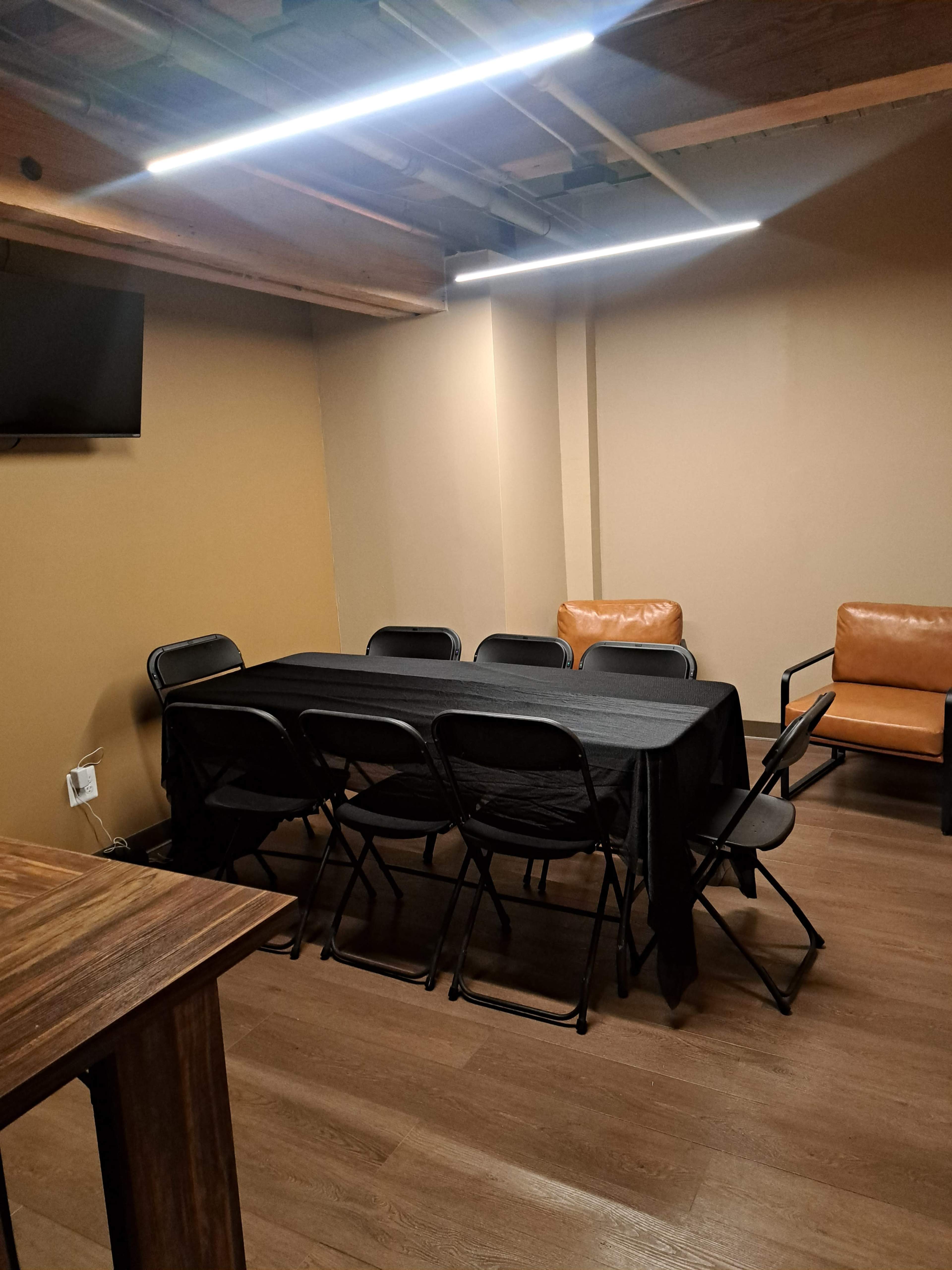 The image shows a small meeting room with a rectangular table covered by a black cloth, surrounded by black folding chairs, and a brown leather armchair against the wall.