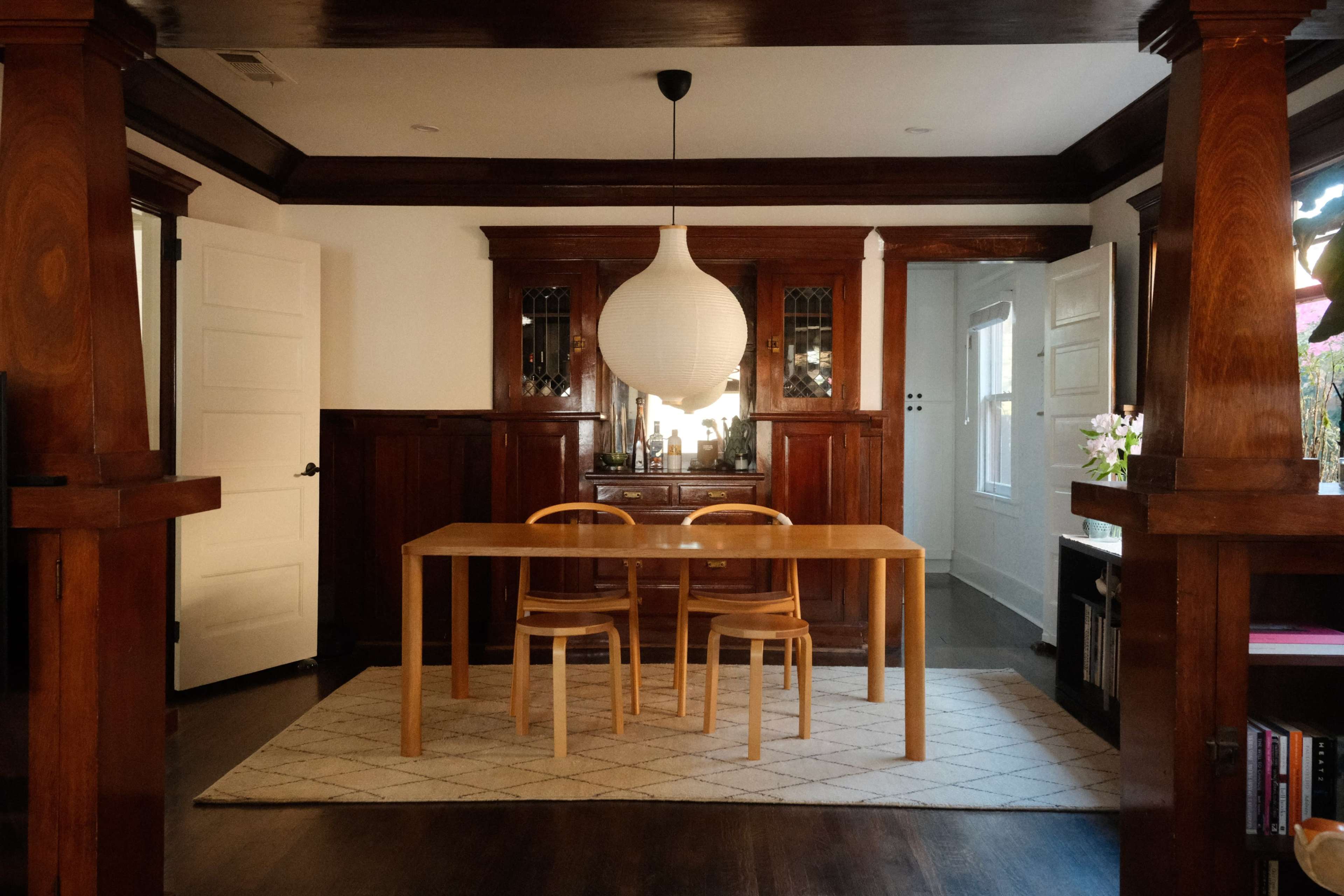 A wooden dining table with two chairs is centered in a room that features dark wooden trim and a large pendant light hanging from the ceiling.